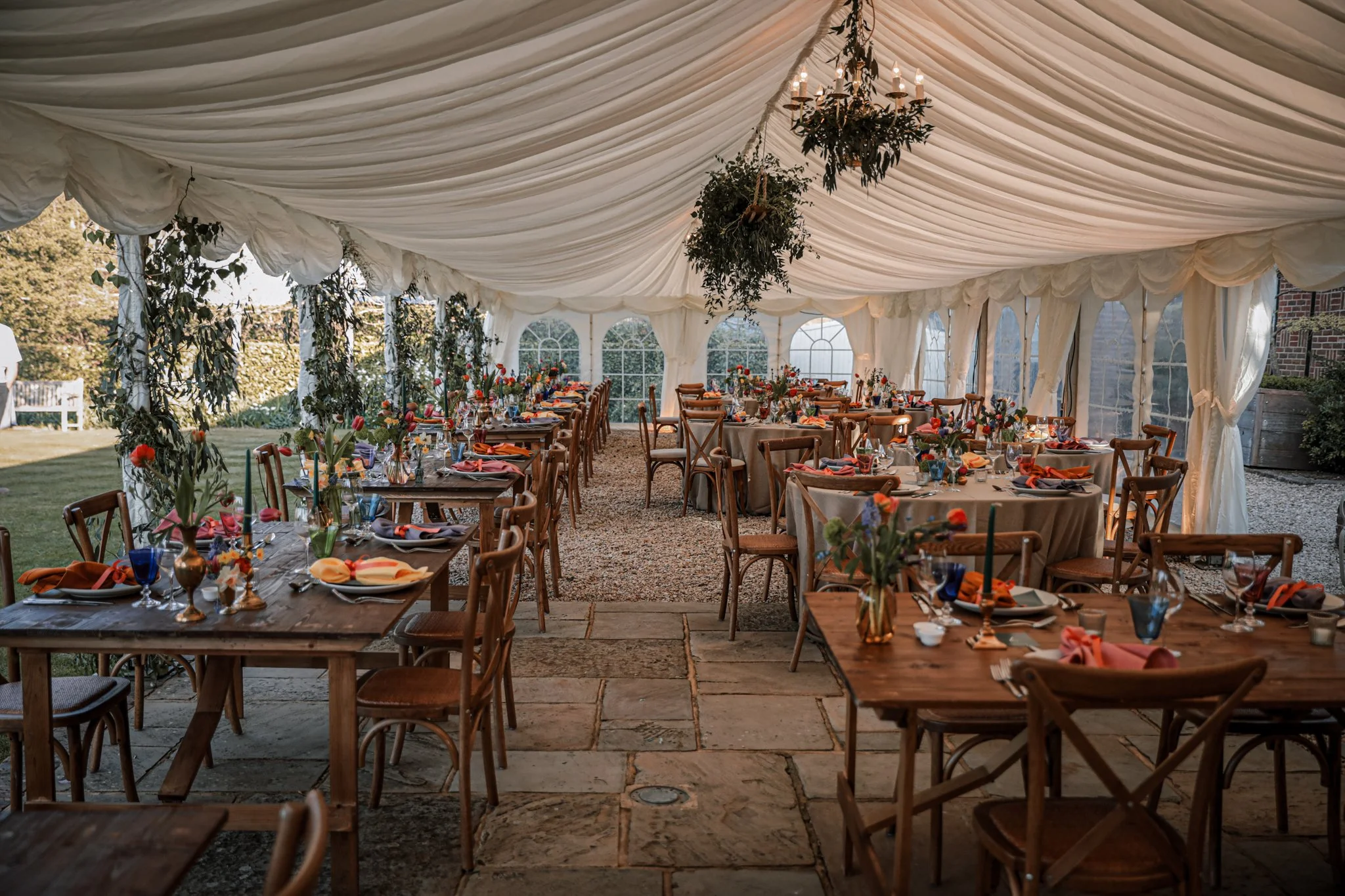 A decorated outdoor event tent with wooden tables, chairs, floral centerpieces, and a chandelier hanging from a draped ceiling.