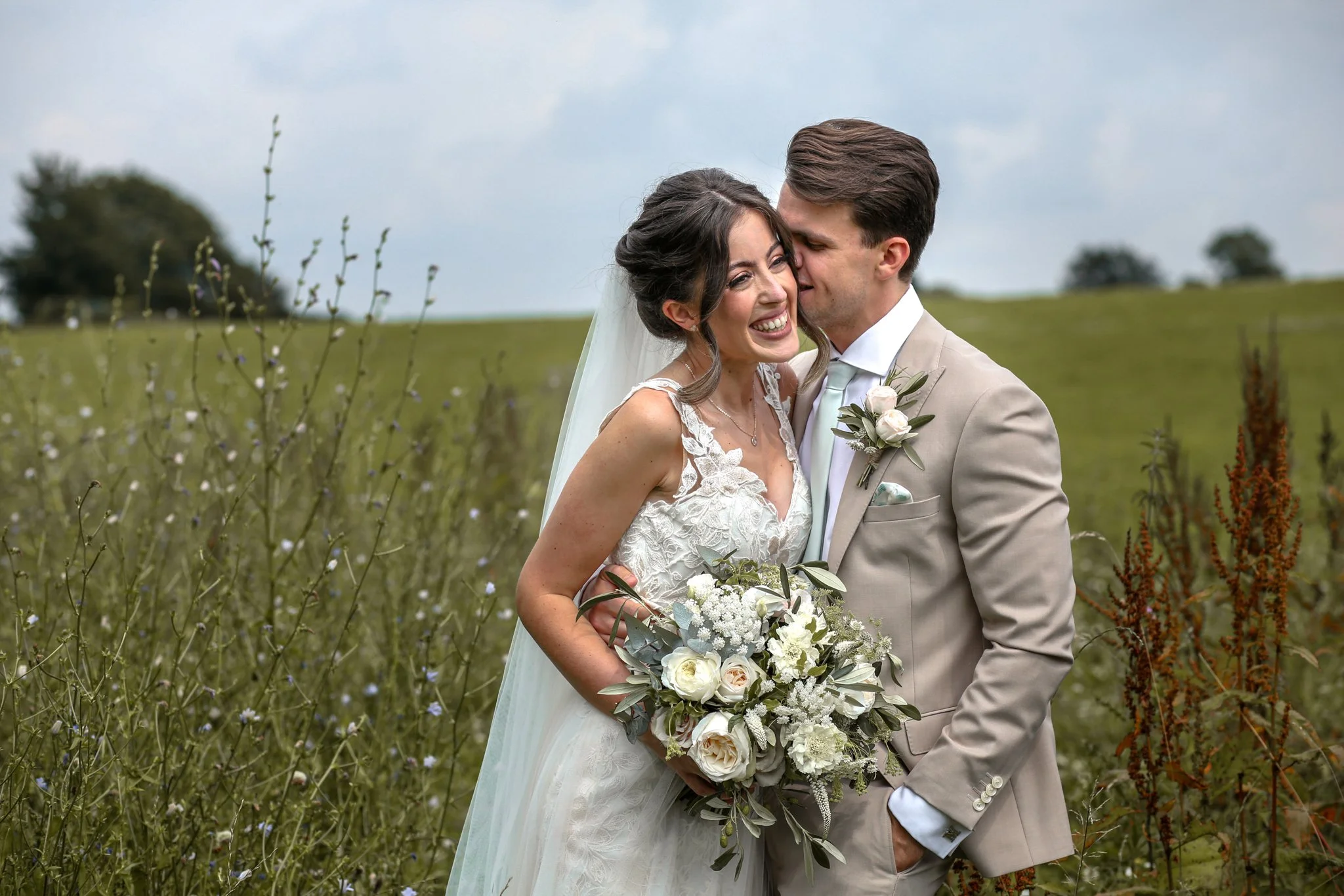 A bride and groom smiling and hugging outdoors in a field with wildflowers, with trees and a cloudy sky in the background.