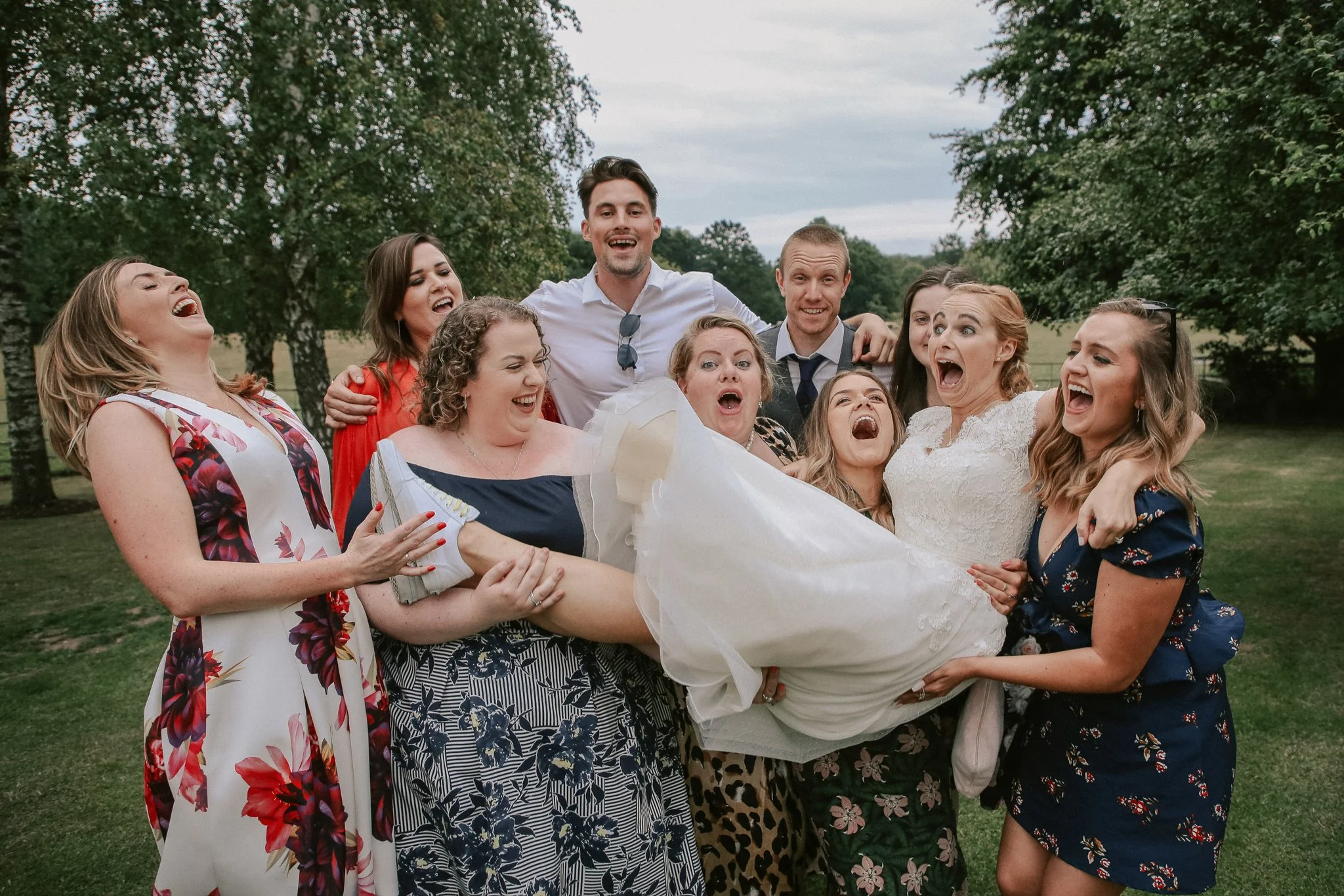 A group of women and two men are outdoors, holding a bride in a white wedding dress who is smiling and appears excited. The group is celebrating, and everyone is laughing or smiling, with trees and an overcast sky in the background.
