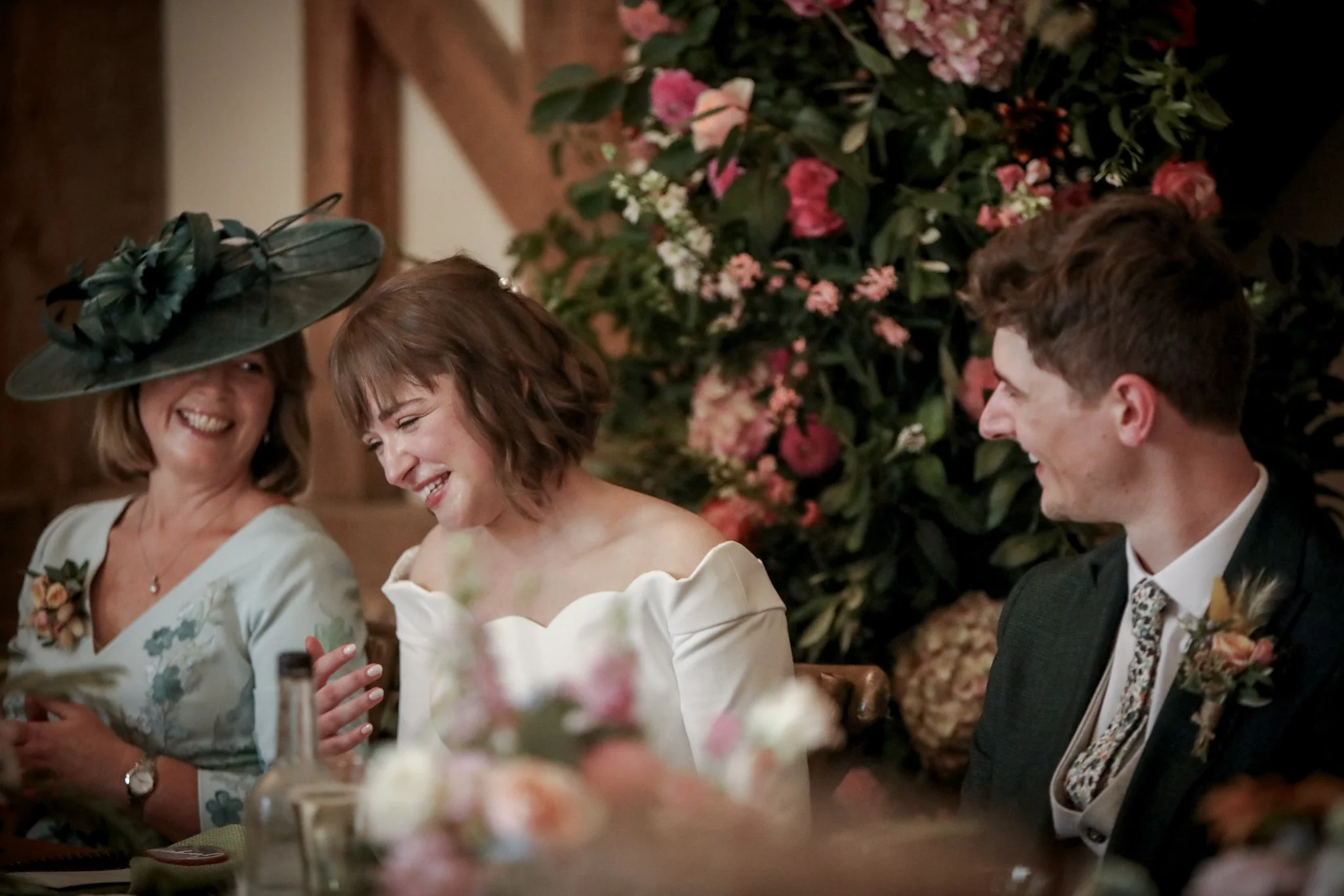 People at a formal event, possibly a wedding, sitting at a table with floral decorations. Three individuals are visible: a woman with a floral hat and a woman with a short haircut holding a smartphone, and a man in a suit and tie. They are smiling an