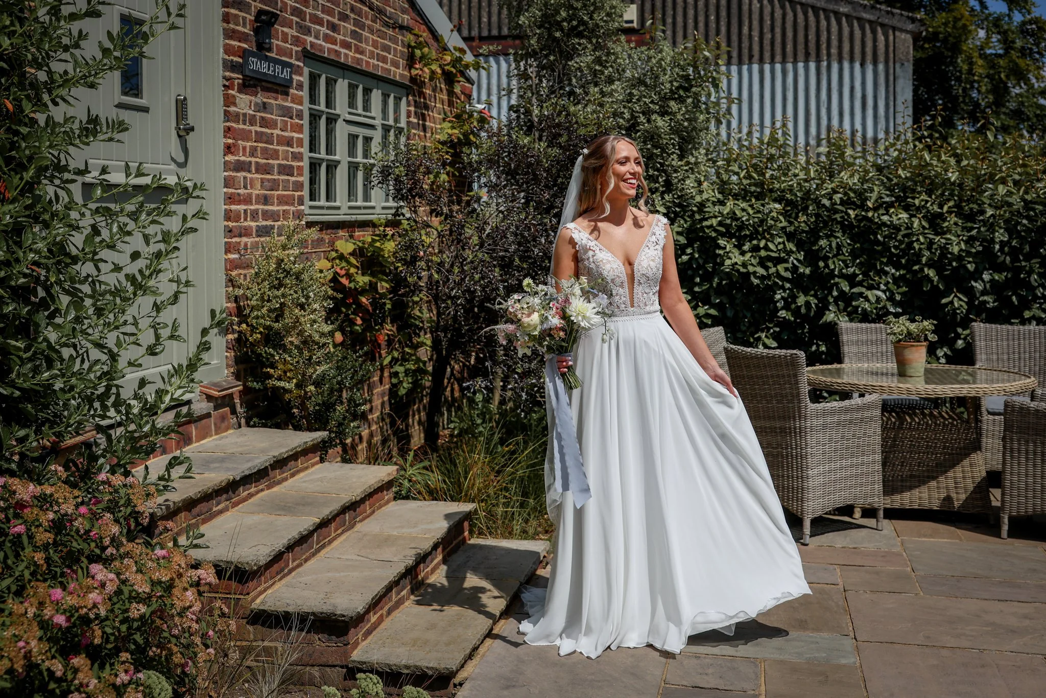 A bride in a white wedding dress holding a bouquet of flowers, smiling outdoors near a brick building and garden.