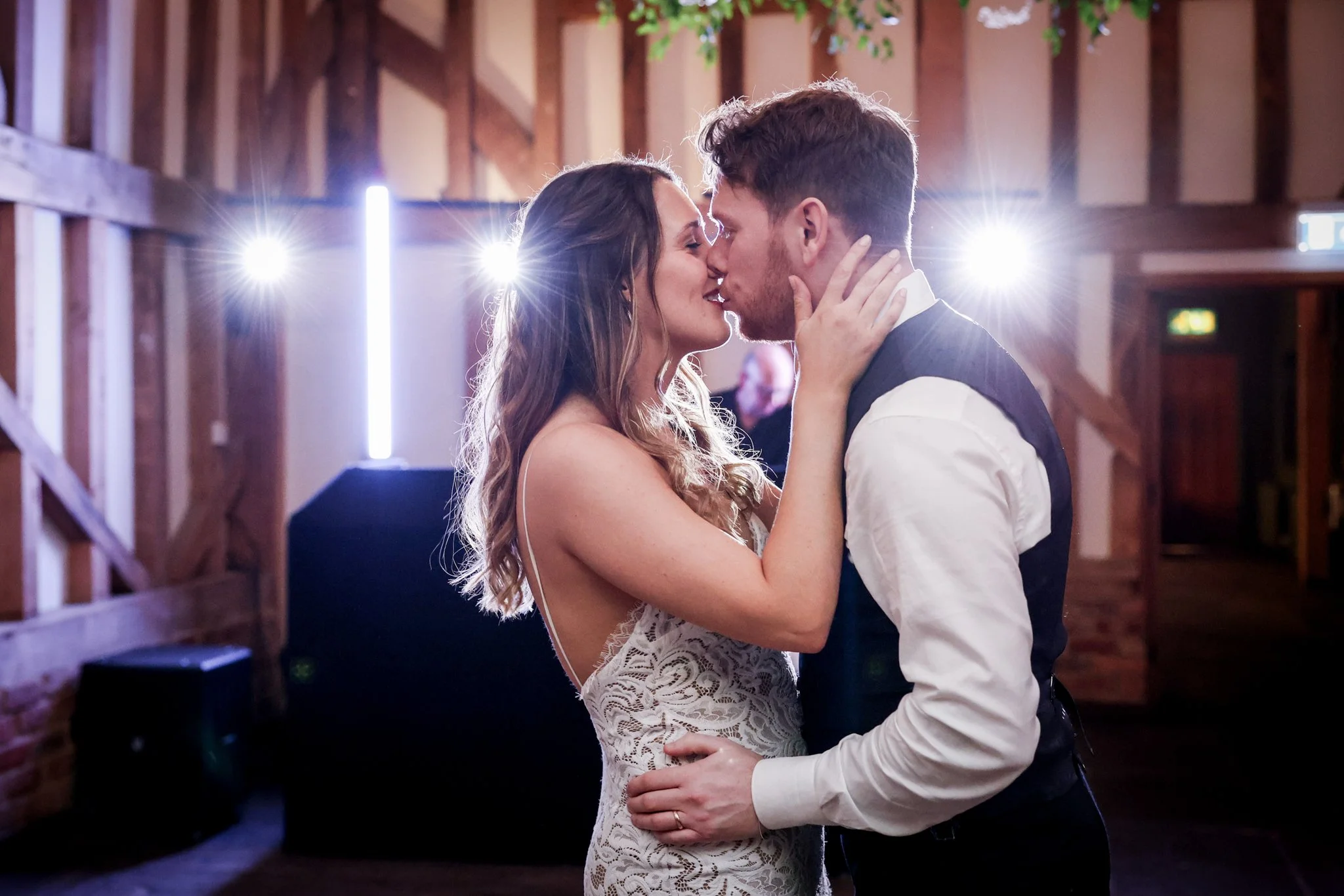 A couple dancing closely in a rustic wooden venue, with the woman wearing a lace dress and the man in a vest and white shirt, illuminated by backlighting.