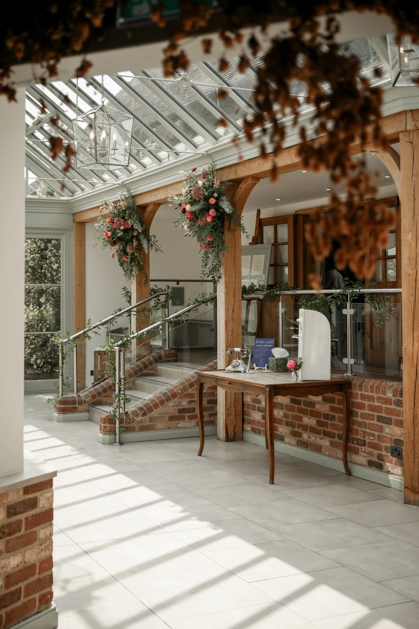 Bright interior with large skylight roof, hanging lantern light, floral arrangements, wooden beams, brick and white wall, and a small wooden table with a sign and flower.