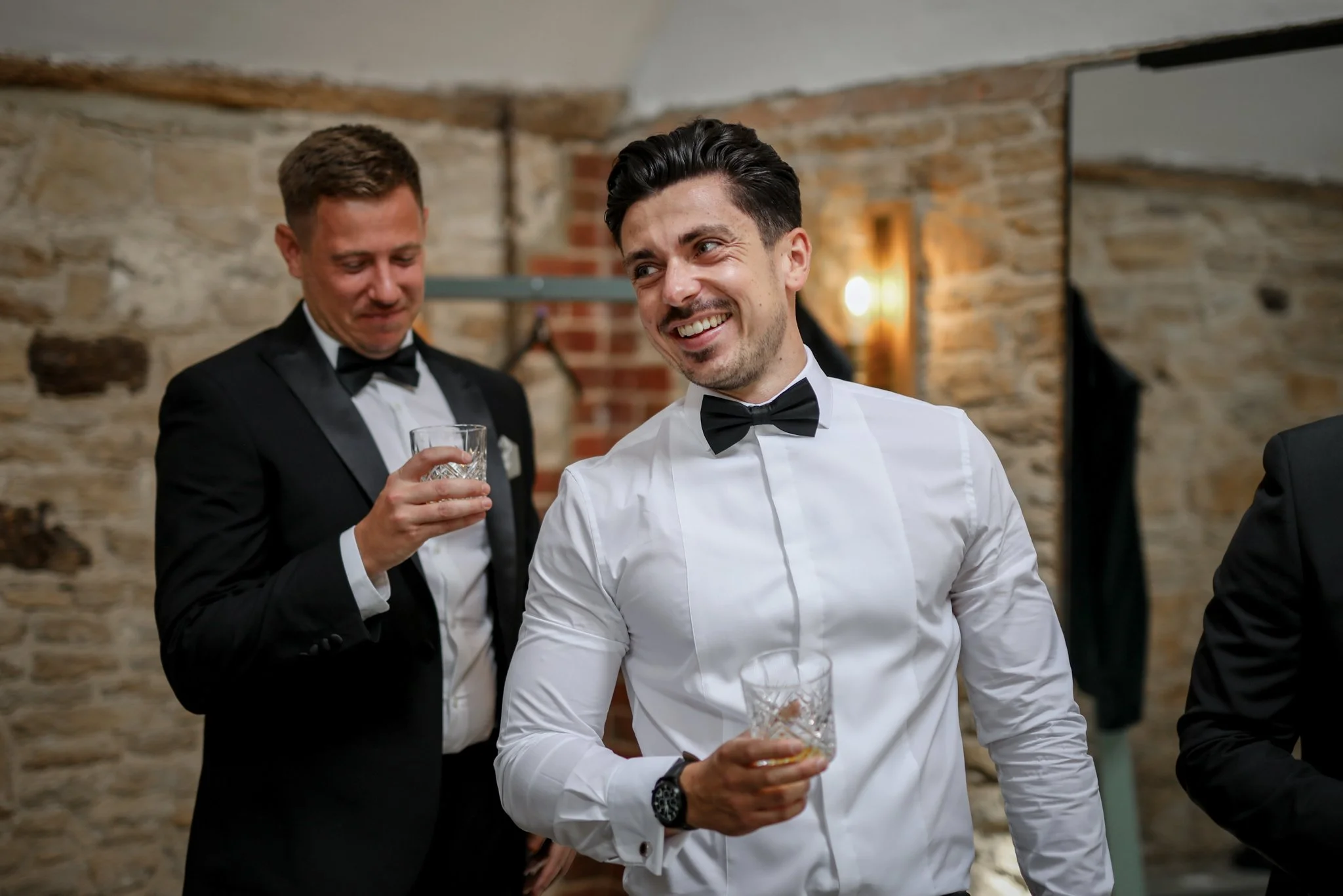 Two men in tuxedos, smiling and holding glasses at a social event in a room with stone walls.