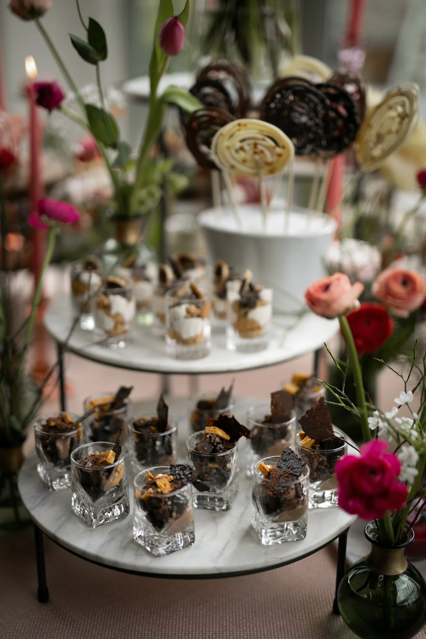 Multiple small glasses filled with desserts on a marble table, with flowers in the background and a large green plant in a vase on the right.