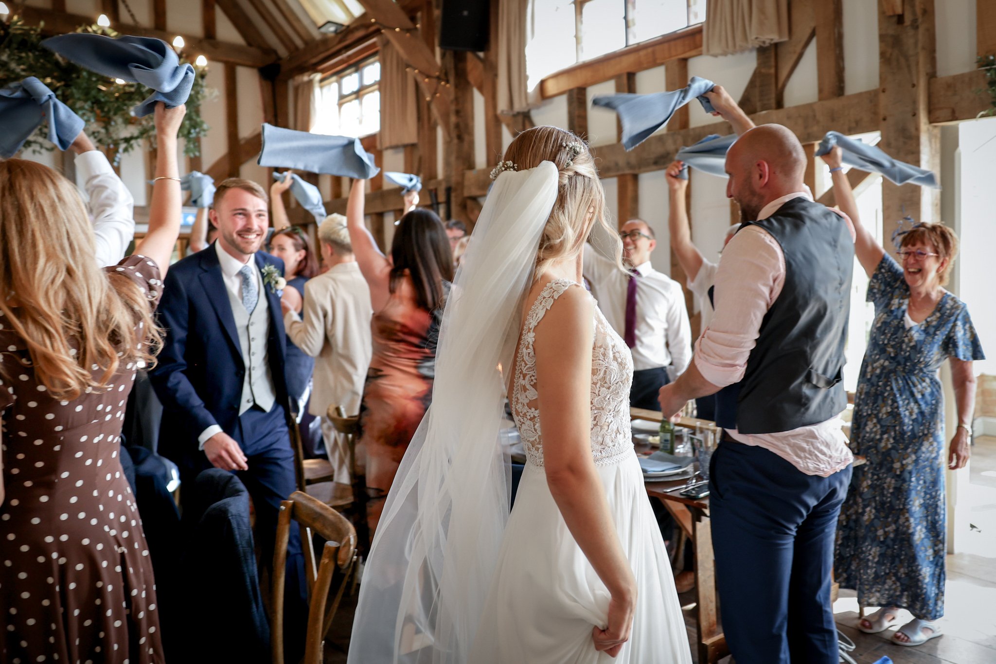 A wedding reception with guests dancing, some waving blue cloths, in a rustic wooden venue. The bride and groom are standing close together, with the bride in a lace wedding dress and veil, and the groom in a suit with a tie. Guests are smiling and e