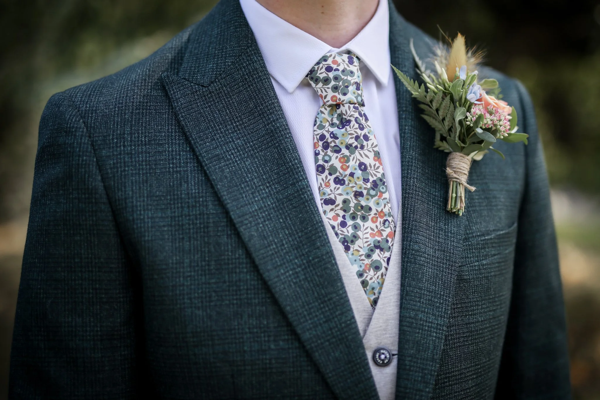 Close-up of a man wearing a dark green checkered suit jacket, white dress shirt, and a floral tie. He has a boutonniere with pink, purple, and white flowers and greenery pinned to his jacket.