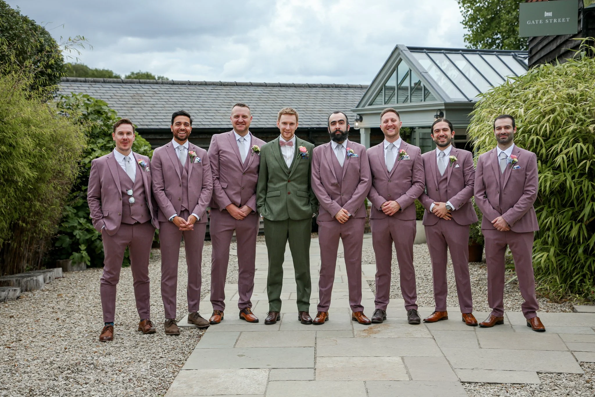 A group of eight men dressed in coordinated suits, with the groom in a green suit and the groomsmen in mauve suits, standing outdoors on a stone pathway and smiling, with a garden and building with a greenhouse roof in the background.