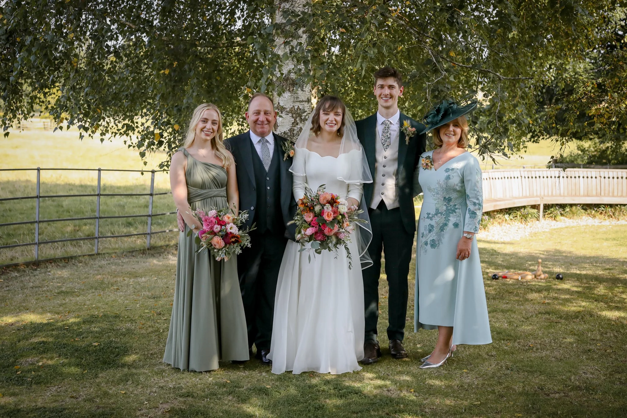 Group of six people standing outdoors beneath a large tree, dressed for a wedding, smiling and holding bouquets, with trees and a grassy area in the background.