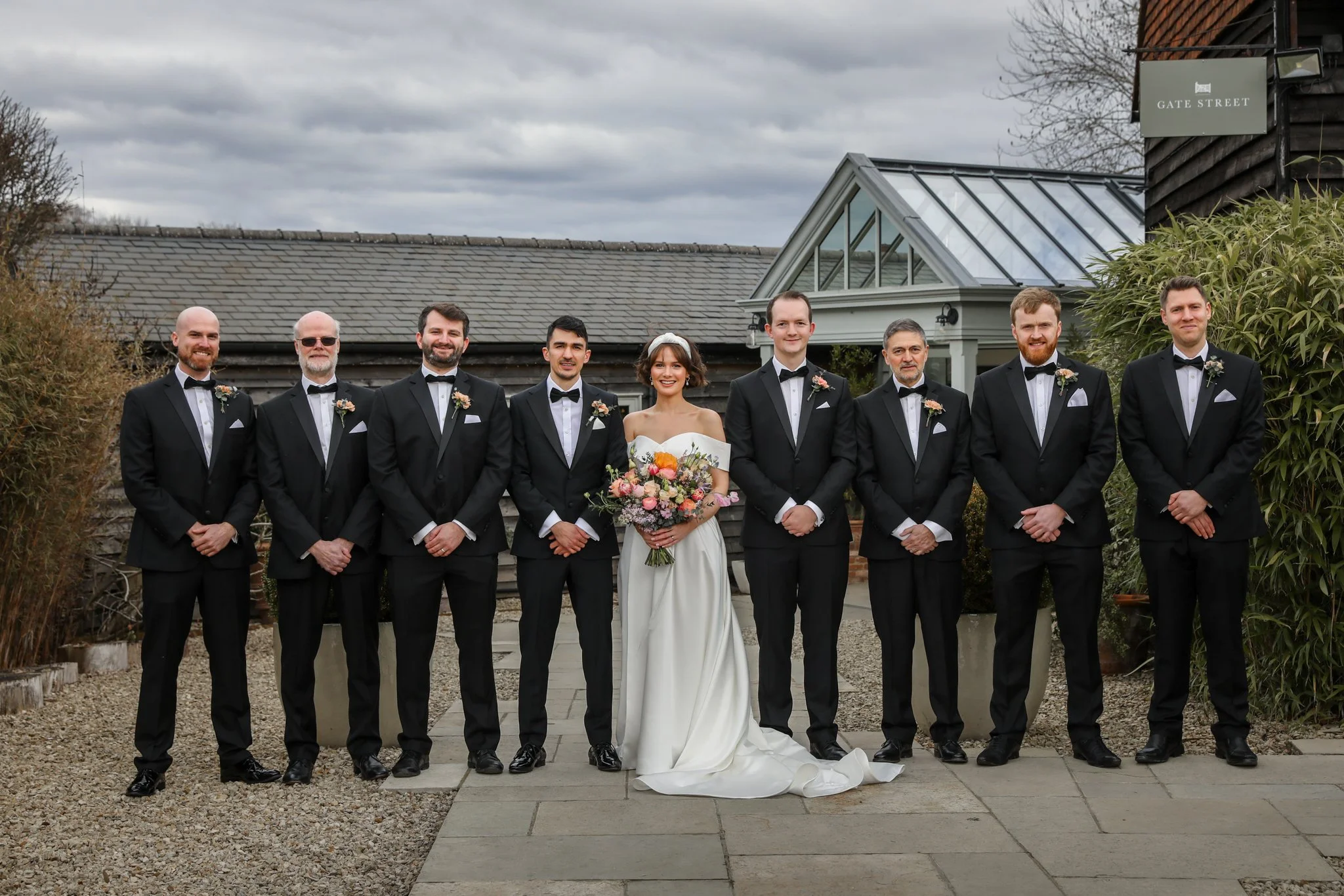 A group of nine people dressed in formal wedding attire, including a bride holding a bouquet of flowers. They are standing outside in front of a building with a sign that reads 'Gate Street' and thunderstorms in the sky.