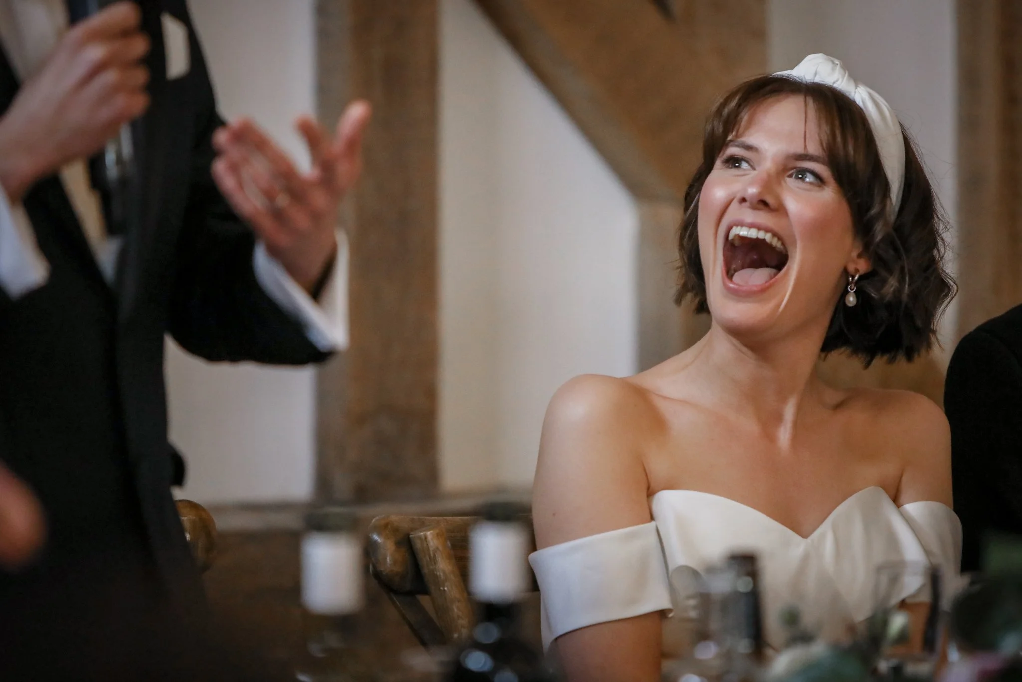 A woman with short brown hair, wearing pearl earrings and a white off-shoulder dress, laughing with her mouth open during a celebration or event. Someone is taking her picture, and part of their hands and a black suit are visible on the left side of 