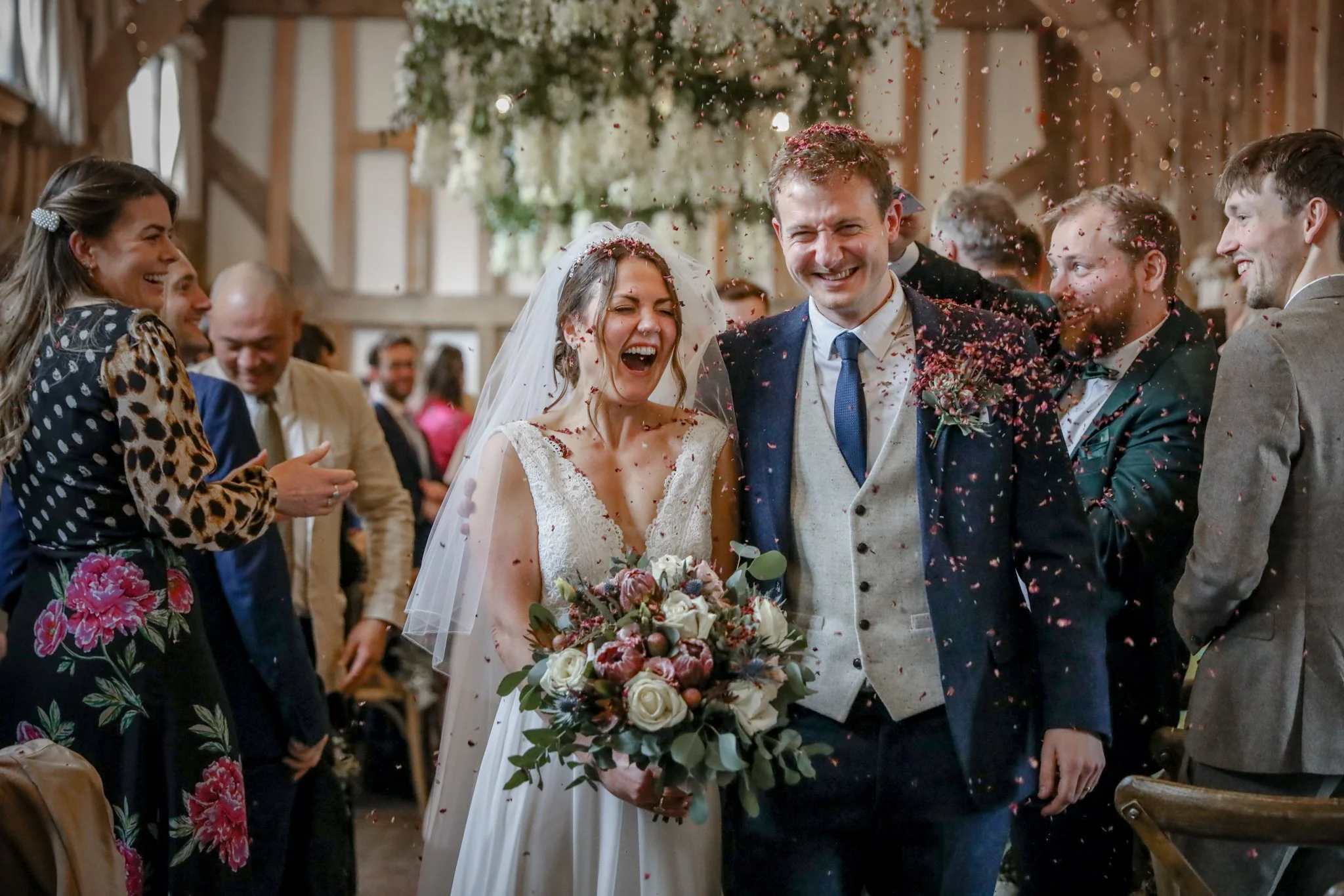 A bride and groom smiling at each other during their wedding celebration surrounded by guests, with flower petals falling around them in a rustic indoor setting.