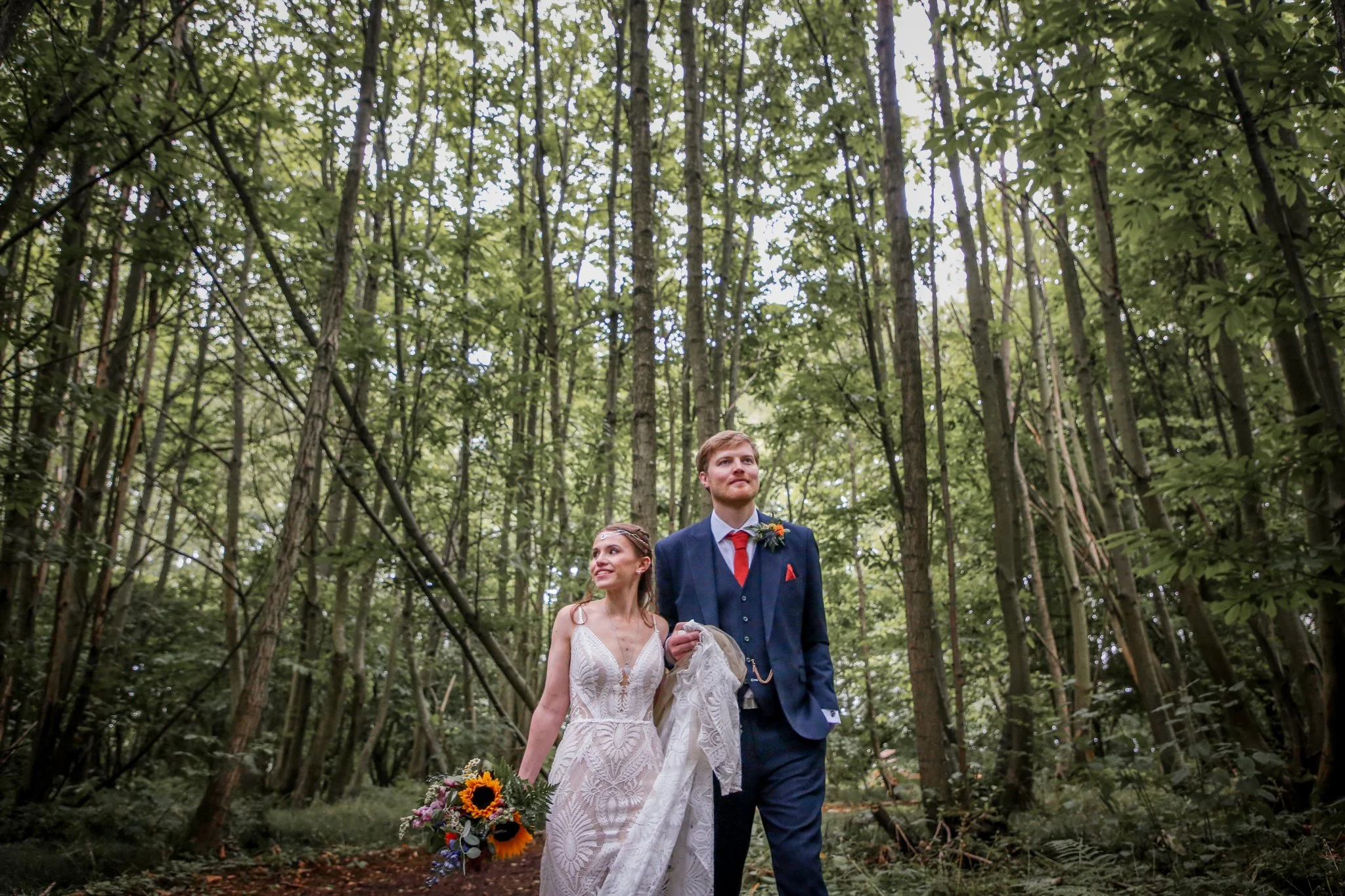 A bride and groom walking together in a forest during their wedding ceremony, with the bride holding a bouquet of sunflowers and the groom wearing a blue suit with a red tie.