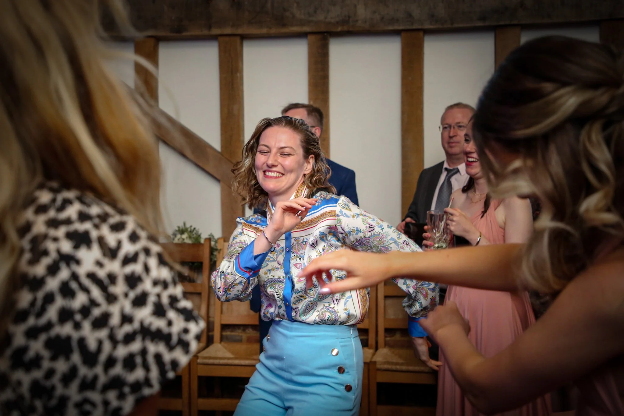 A woman smiling and laughing at a social gathering, surrounded by other people, with wooden stairs in the background.