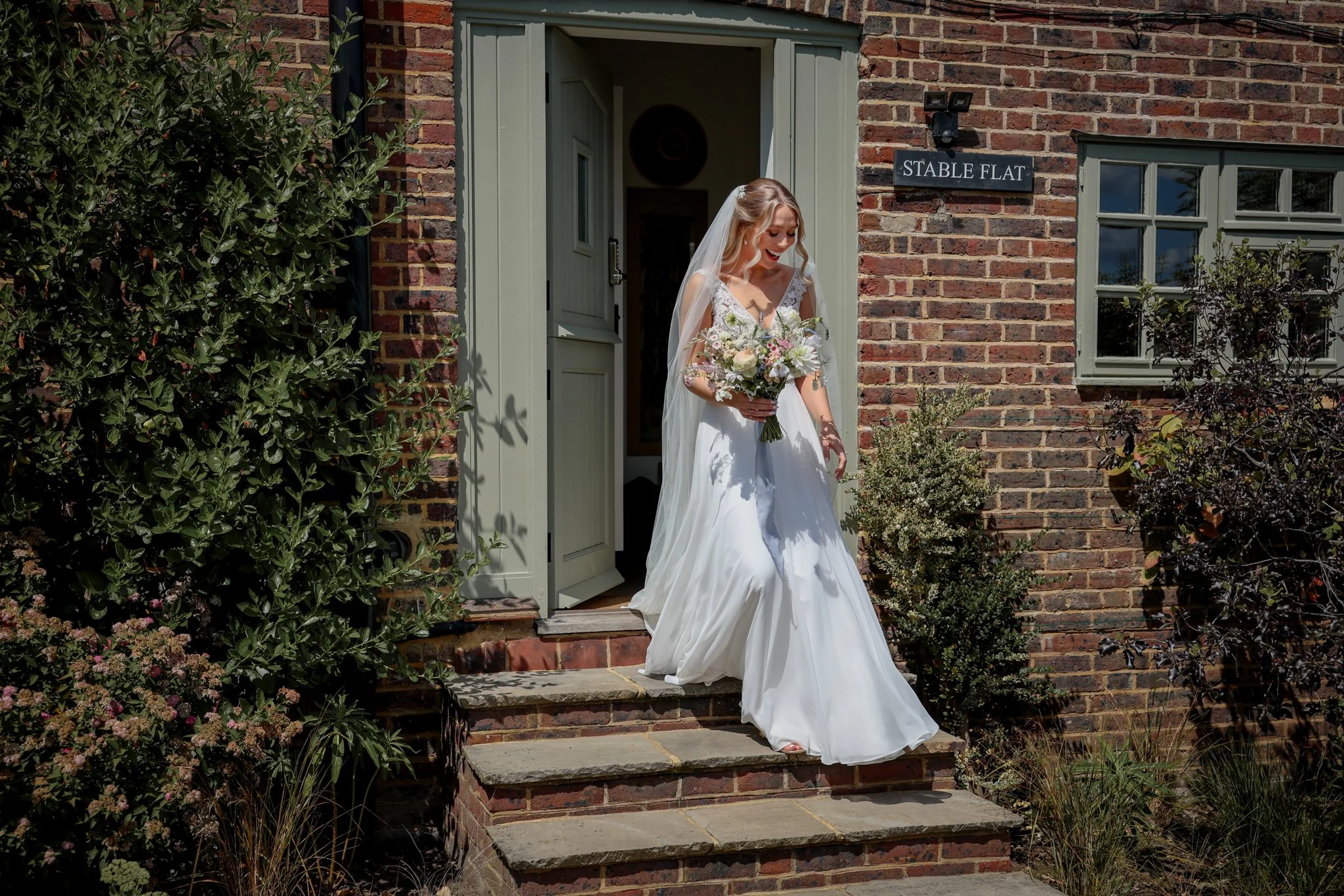 A bride in a white wedding dress, holding a bouquet of flowers, smiling as she descends the brick stairs outside a building labeled "Stable Flat" on a sign on the brick wall.