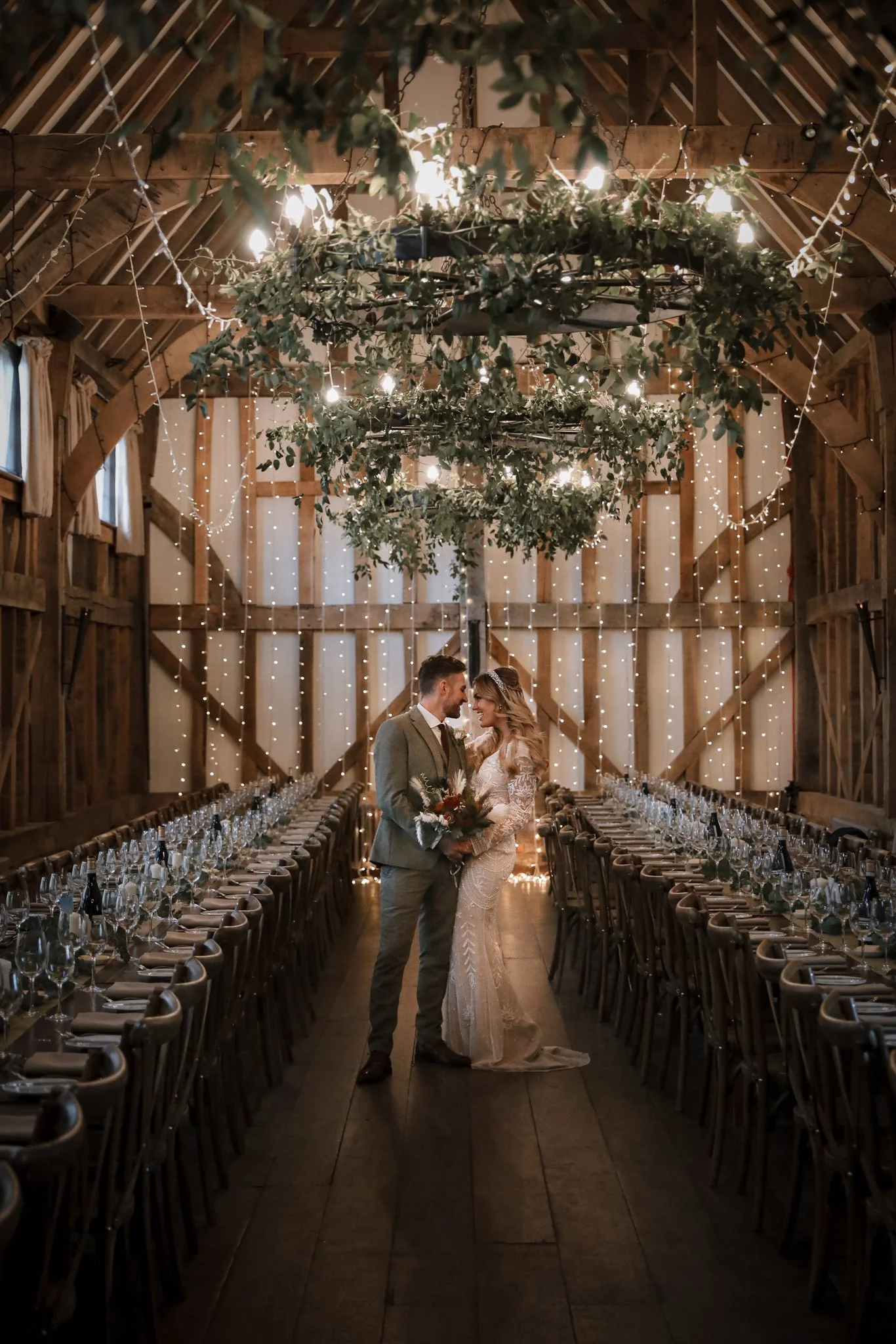 A bride and groom standing close together in the center of a rustic wedding reception hall, decorated with string lights, greenery, and long banquet tables set with glassware and silverware.
