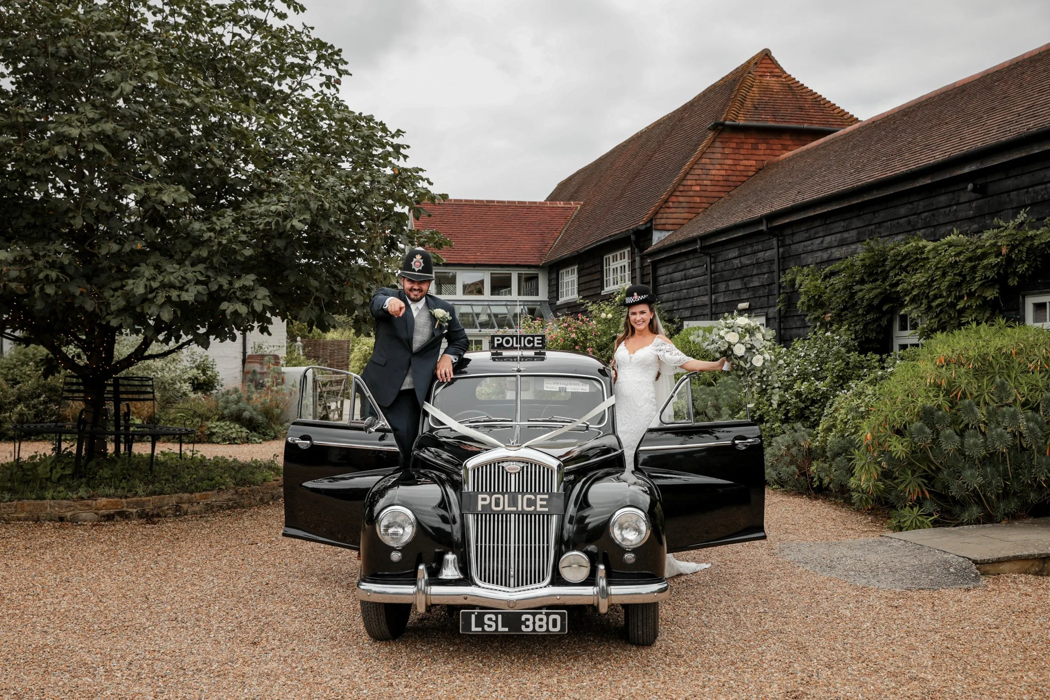 A wedding couple in police costumes posing on a vintage police car in a garden.