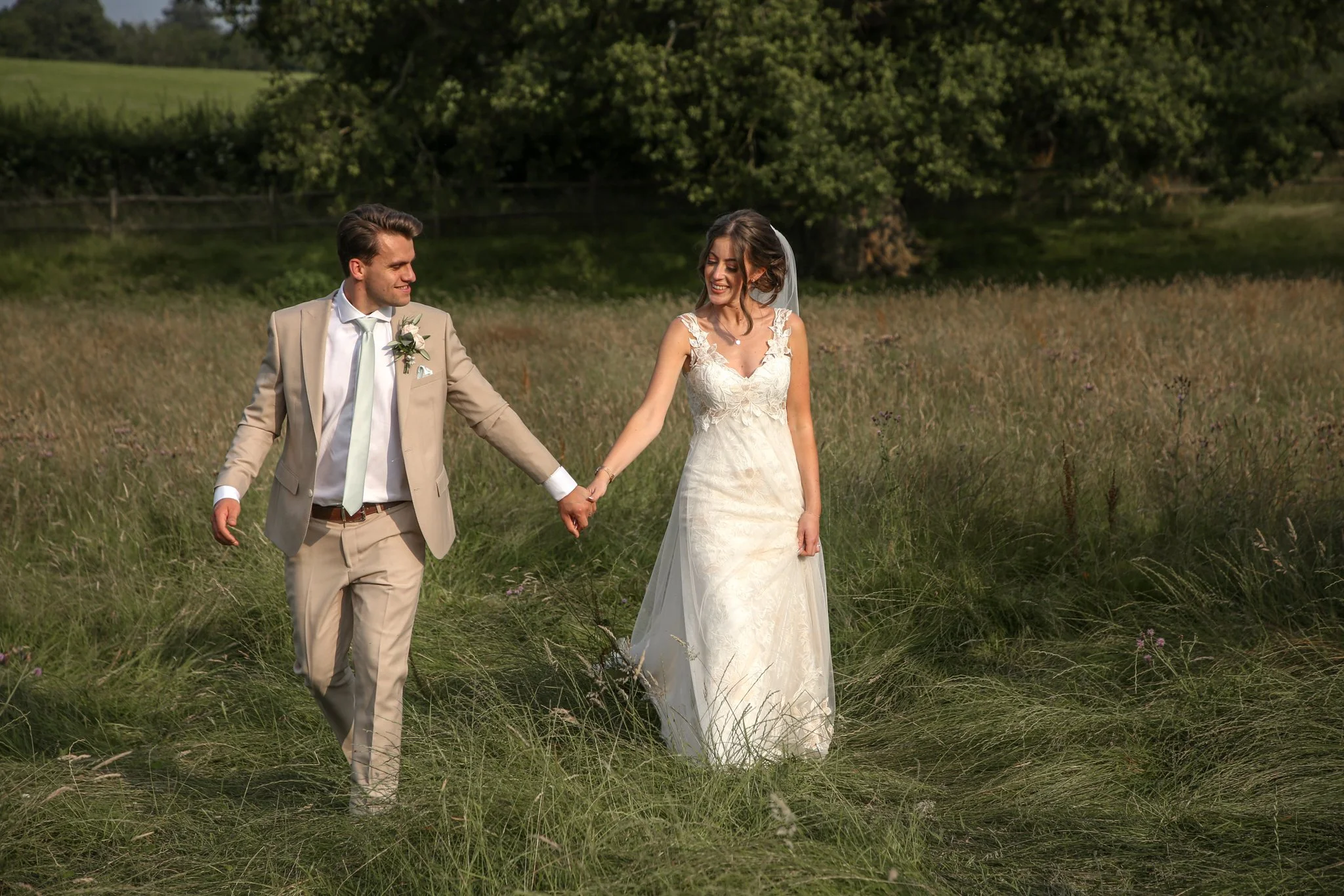 A bride and groom walking hand in hand through a grassy field with trees in the background on a sunny day.