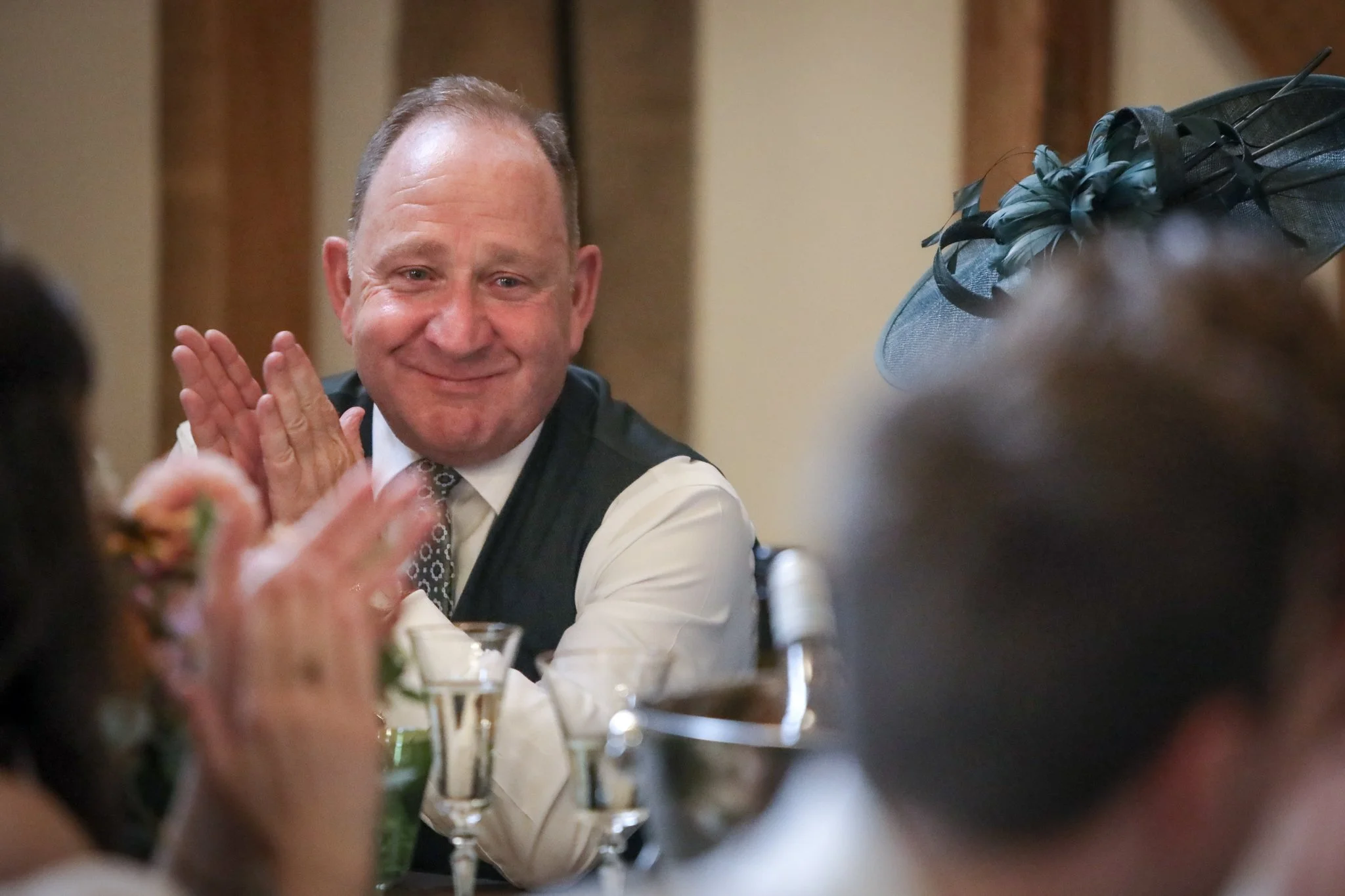 A middle-aged man with short hair, wearing a white shirt and patterned tie, smiling and clapping at a formal event or gathering. There is a woman wearing a dark fascinator with feathers and netting in the foreground, slightly out of focus.
