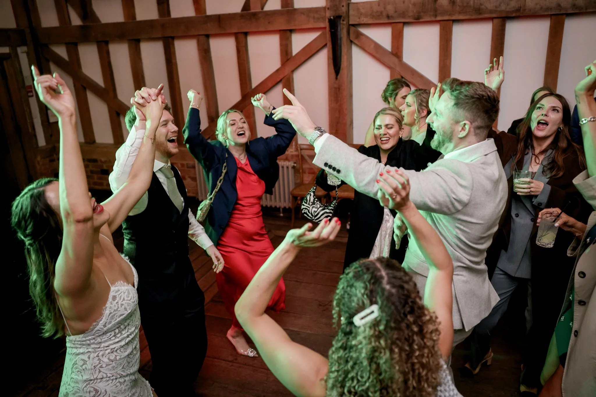 People dancing and celebrating at a party in a wooden duplex or barn setting, with some holding drinks and raising their hands.
