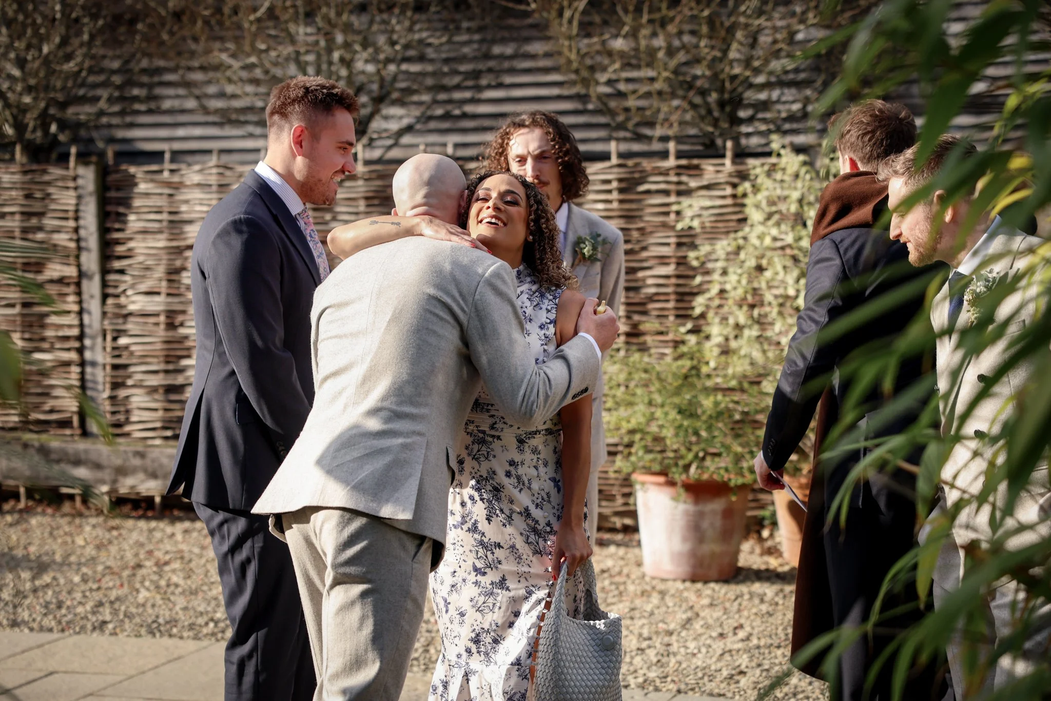 A woman in a floral dress and a man in a suit are hugging a person dressed in a beige suit at an outdoor event, with other well-dressed people nearby and a wooden fence in the background.
