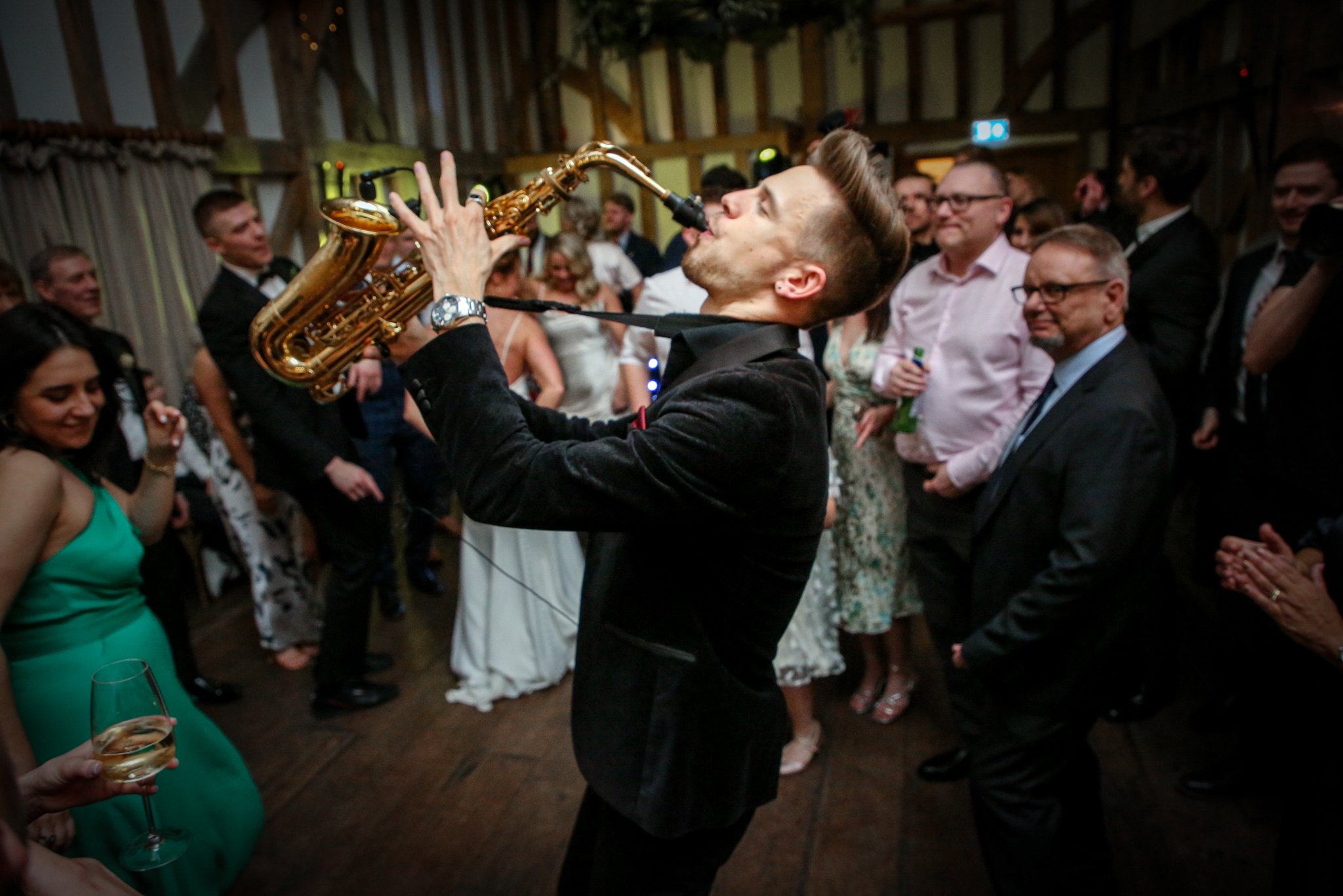 A man in a black tuxedo is playing the saxophone at a party or wedding reception, surrounded by guests who are watching and smiling.