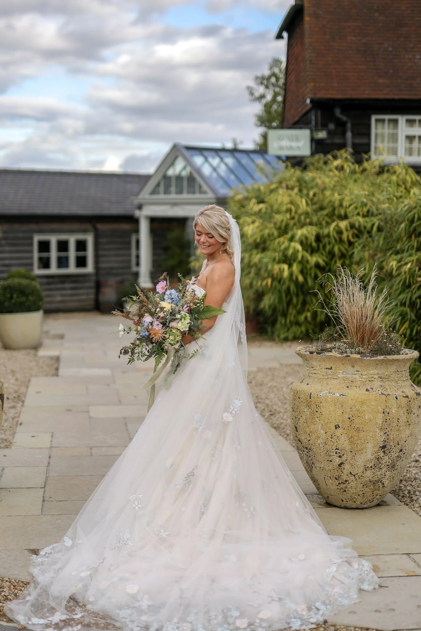Smiling bride in a white wedding dress holding a bouquet of flowers outside near a large decorative plant pot, with a rustic building and cloudy sky in the background.