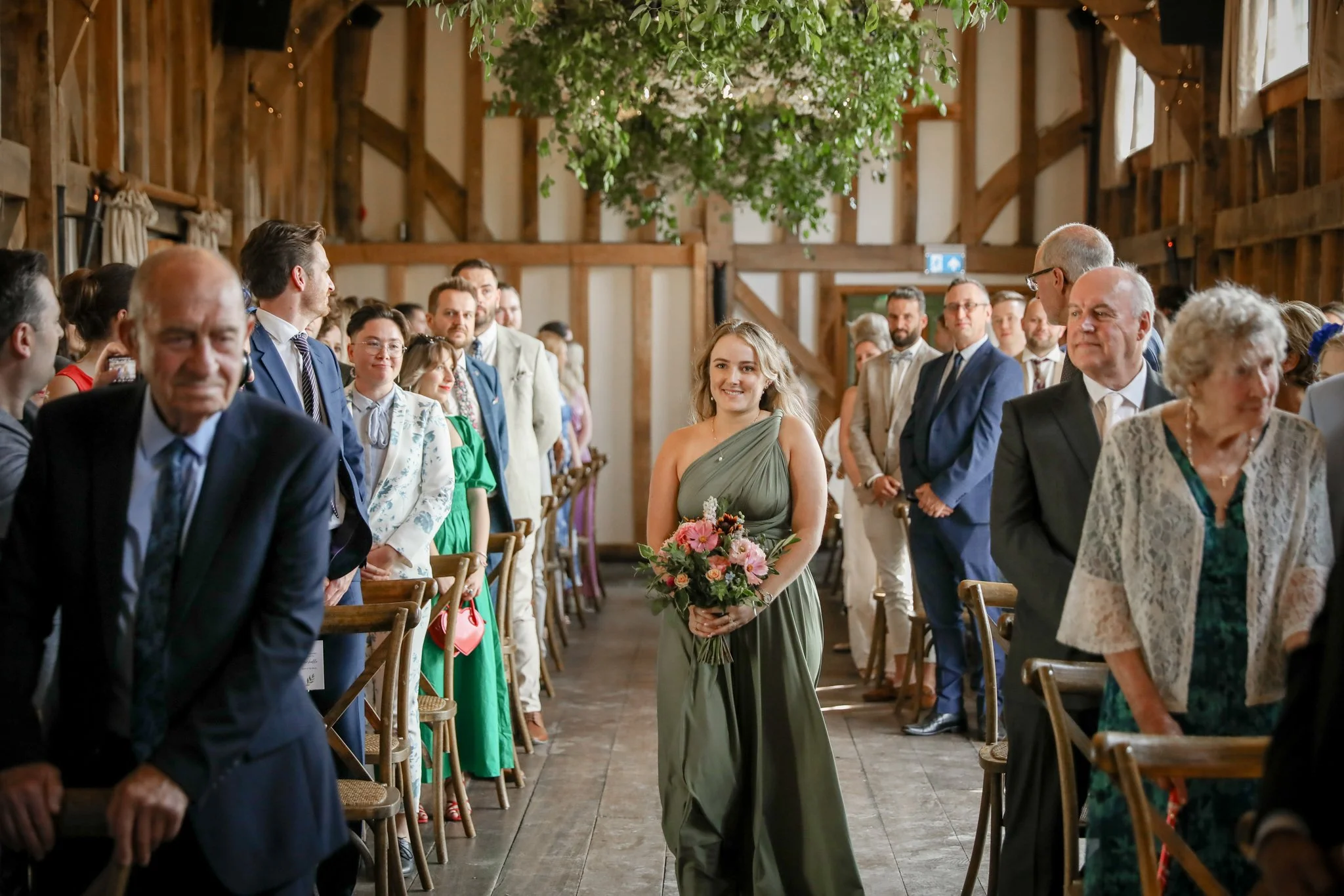 Guests seated and standing in a rustic barn wedding ceremony, with a smiling woman in a green dress holding a bouquet of pink flowers, surrounded by wooden chairs and green hanging plants.