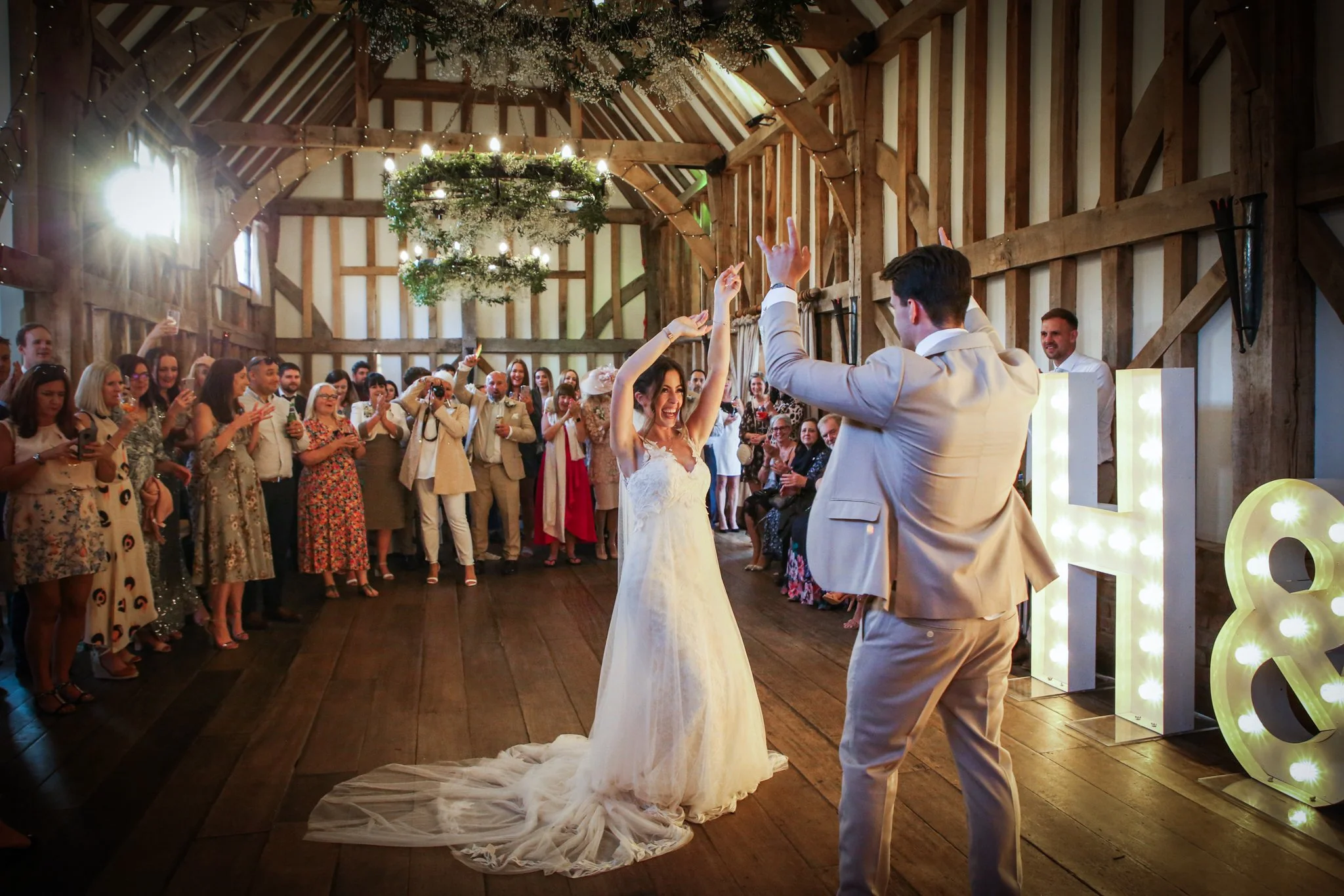 A bride and groom dancing at their wedding reception in a rustic barn with many guests watching and smiling.