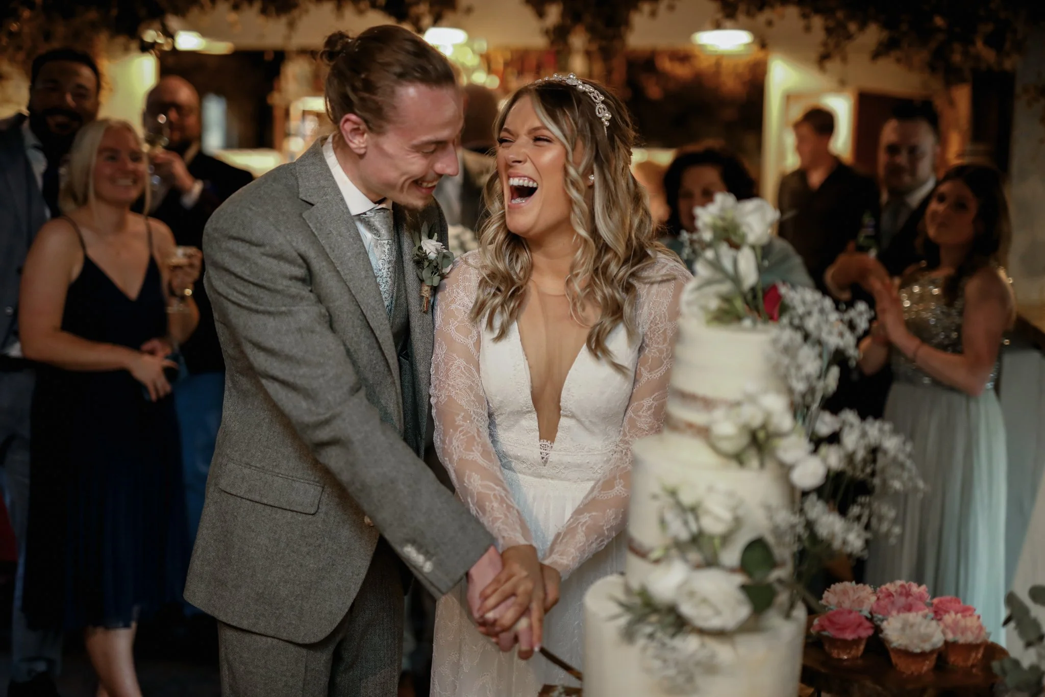 A bride and groom smiling and holding hands during their wedding reception, surrounded by friends and family, with a cake decorated with flowers in front of them.