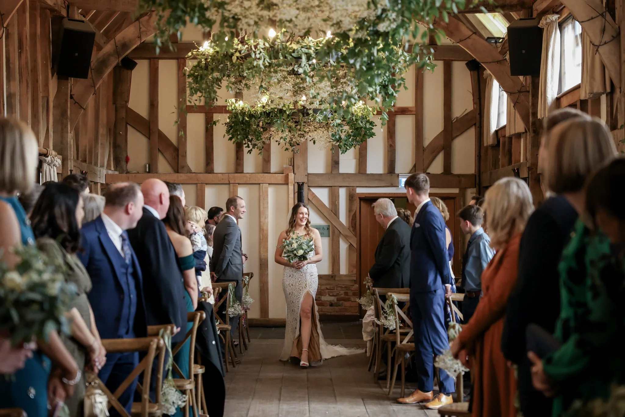 A wedding ceremony in a rustic barn with wooden walls and ceiling, decorated with hanging greenery and flowers, featuring a bride walking down the aisle surrounded by seated guests.