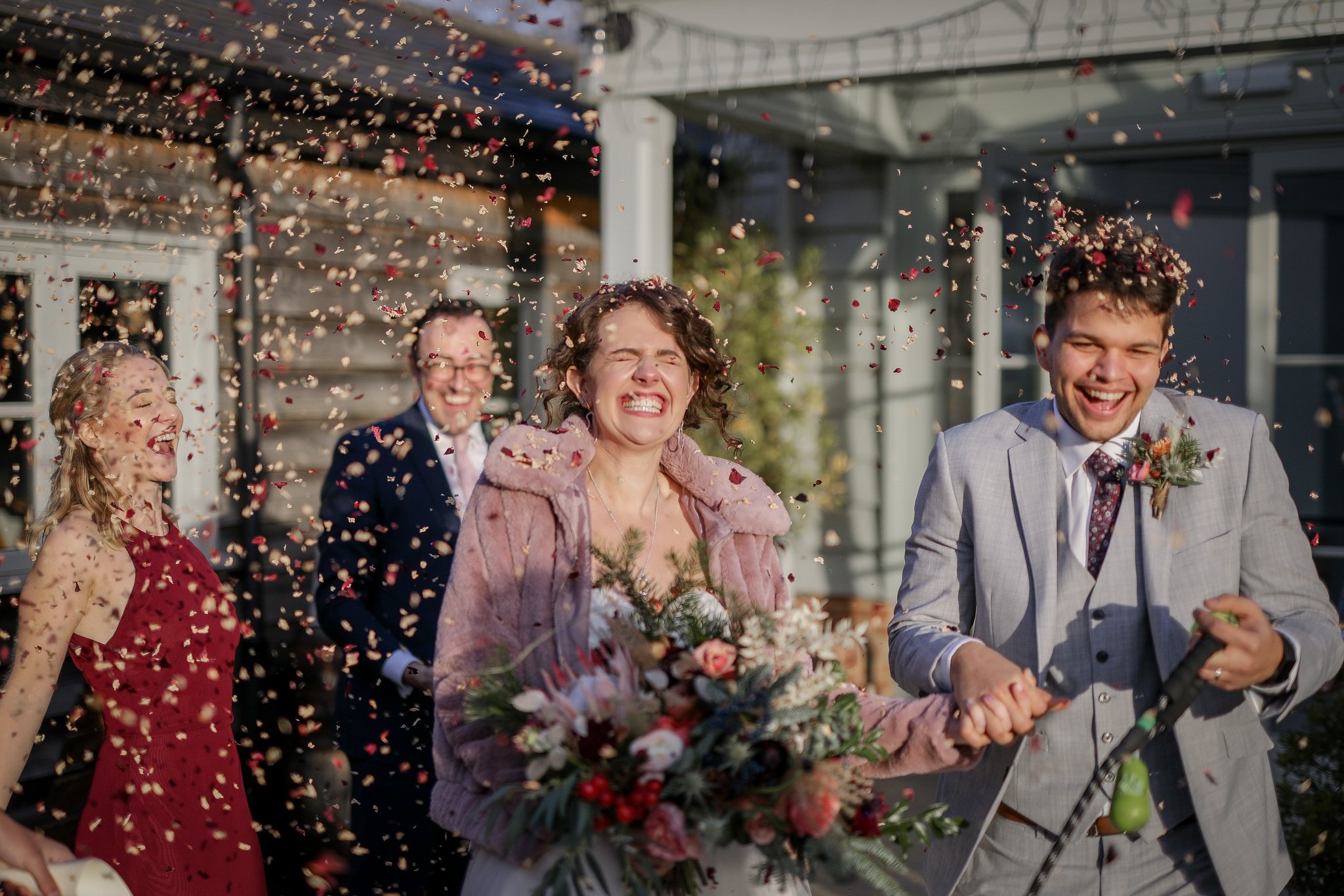 A group of people at a wedding, throwing flower petals while smiling and enjoying the celebration outdoors.