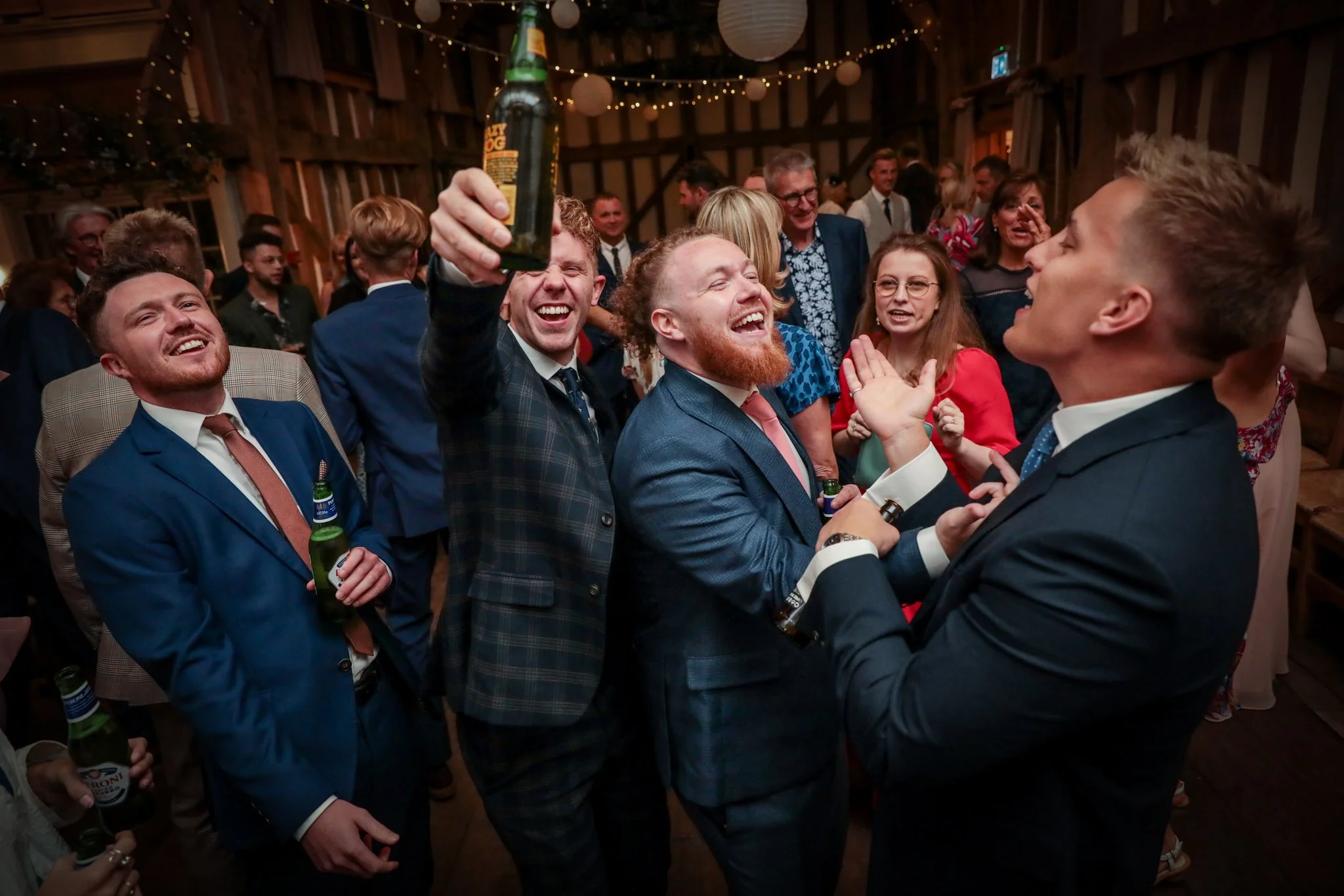 Group of people at a lively celebration or party, many smiling and holding drinks, in a decorated rustic venue with hanging paper lanterns and string lights.