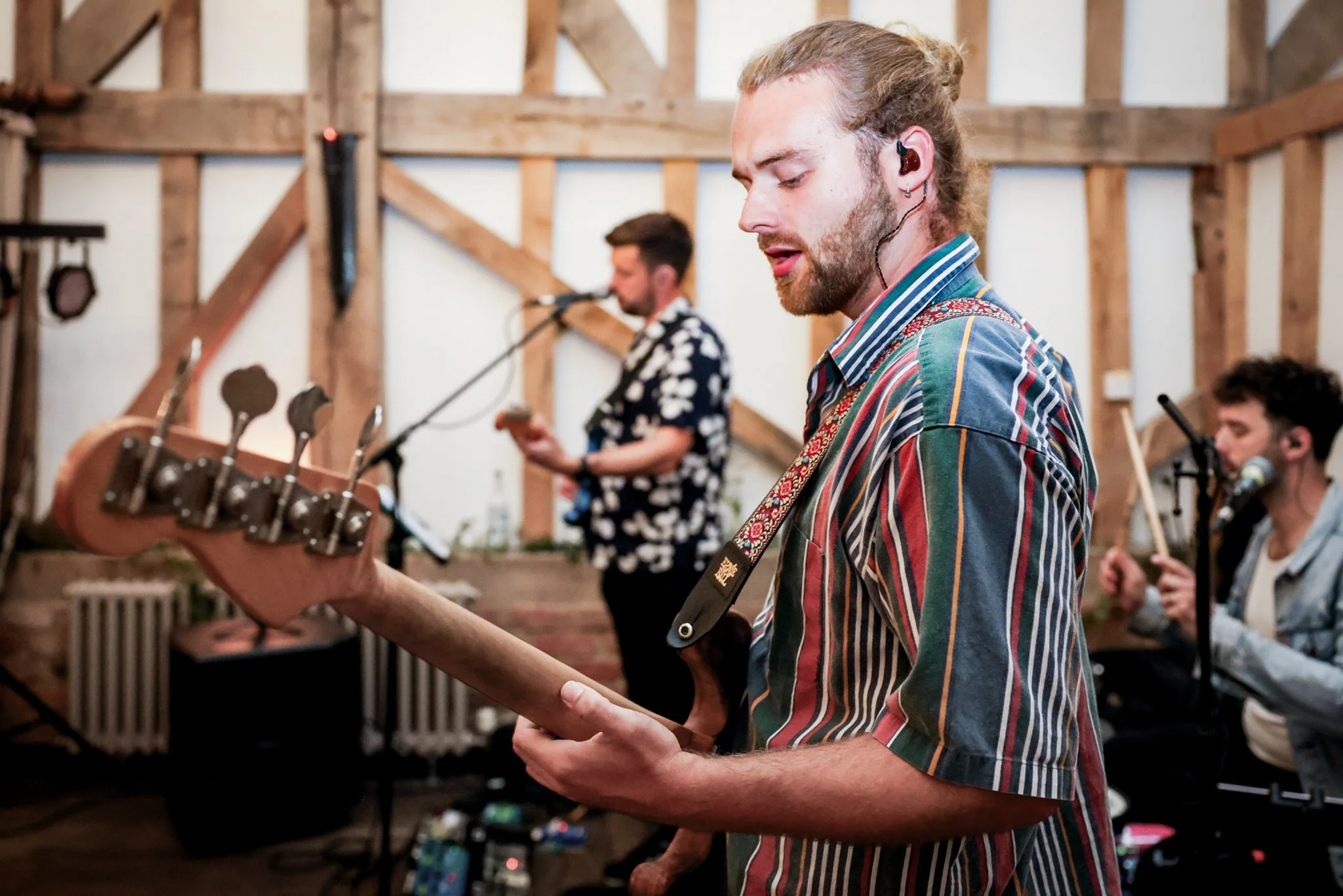 A young man with long hair and a beard playing electric guitar at a music session in a rustic wooden room. Other musicians are visible in the background.