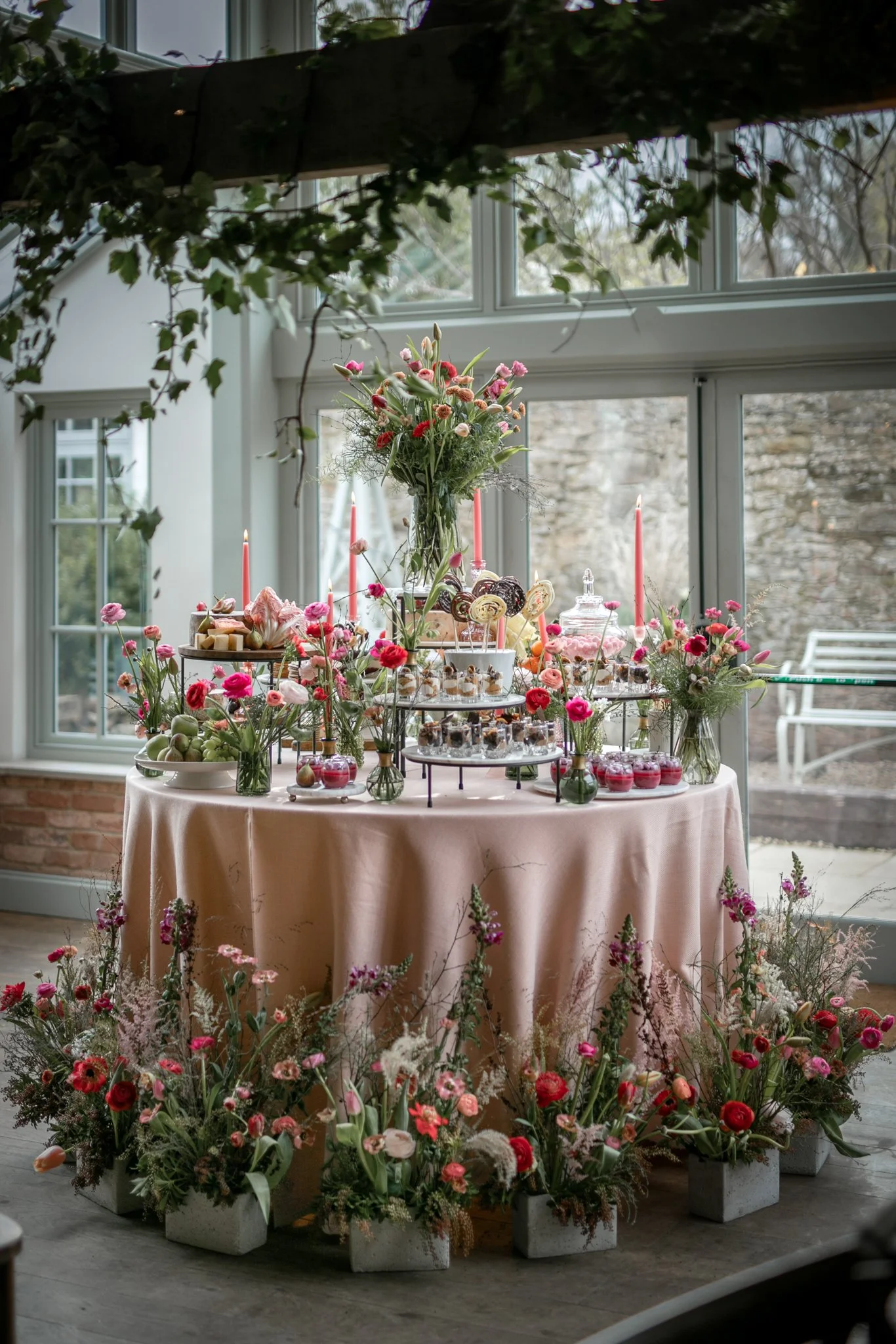 Table decorated with a pink tablecloth, filled with pink and red flowers, sweets, and desserts. Tall flower arrangements, pink candles, and various treats. Large windows in the background with natural light.