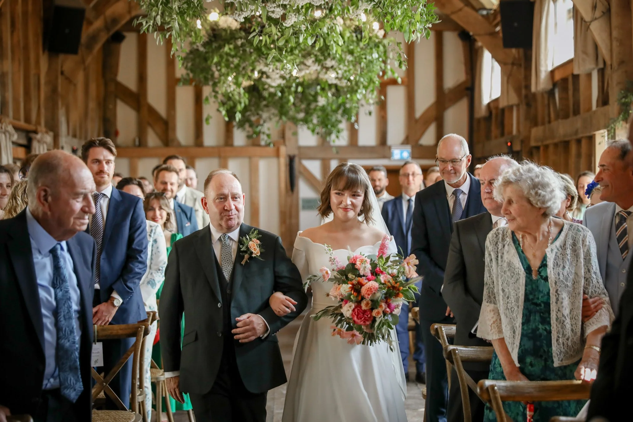 A bride and groom standing together at a wedding ceremony inside a rustic barn with wooden walls and hanging green foliage, surrounded by seated guests.