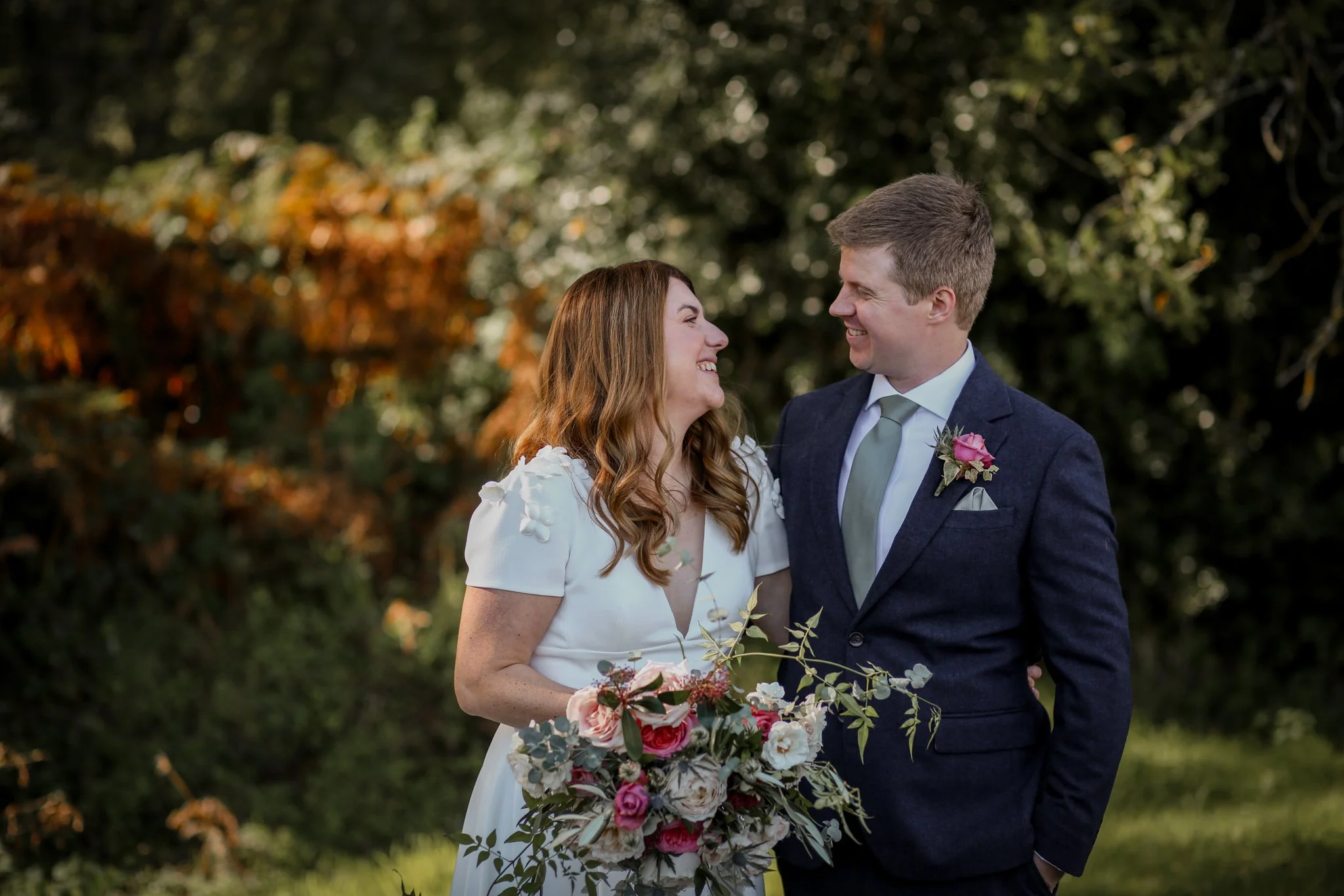 A bride and groom smiling at each other outdoors during a wedding, with trees and greenery in the background.