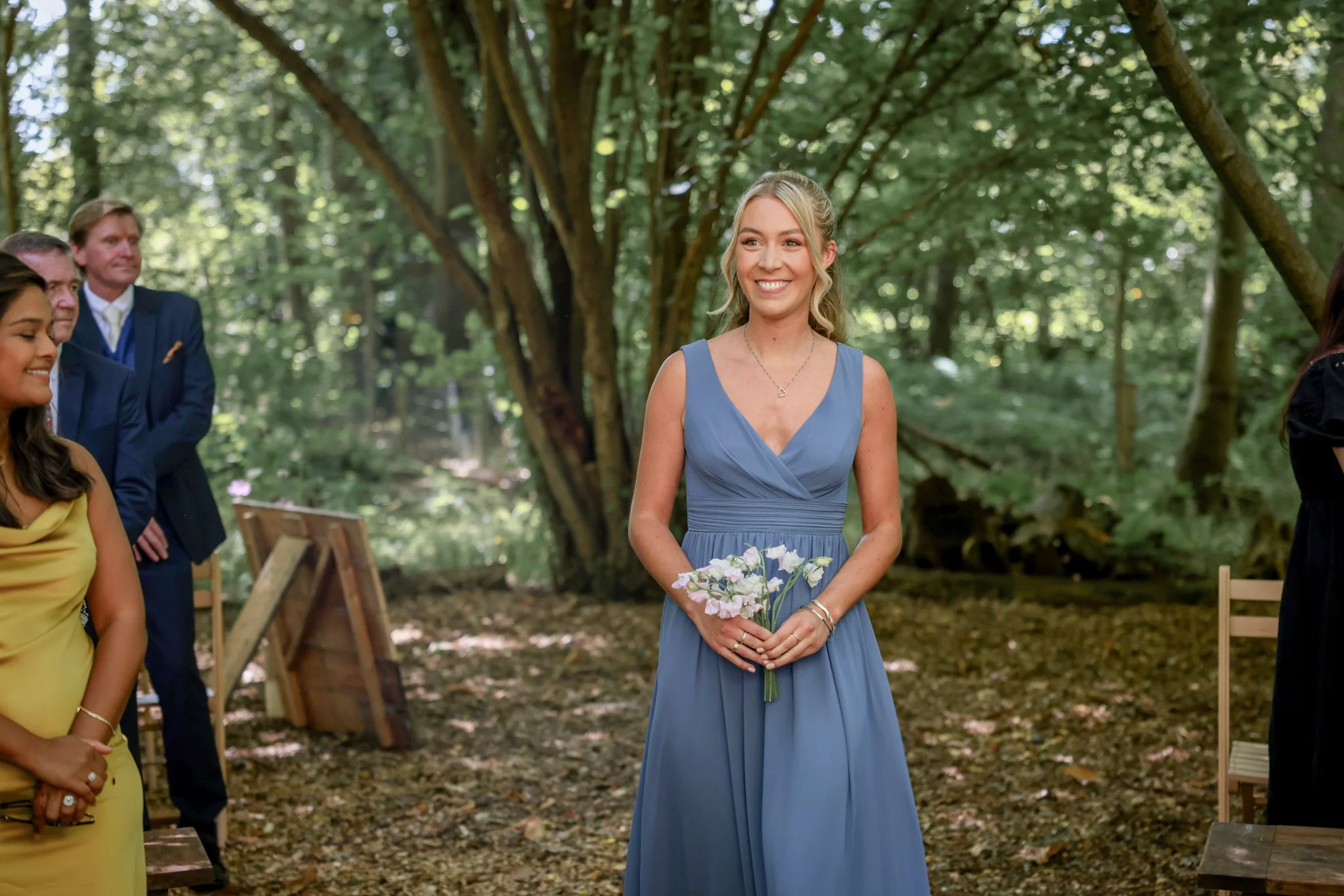 A smiling woman in a blue dress holding a bouquet of flowers at an outdoor wedding ceremony surrounded by trees.