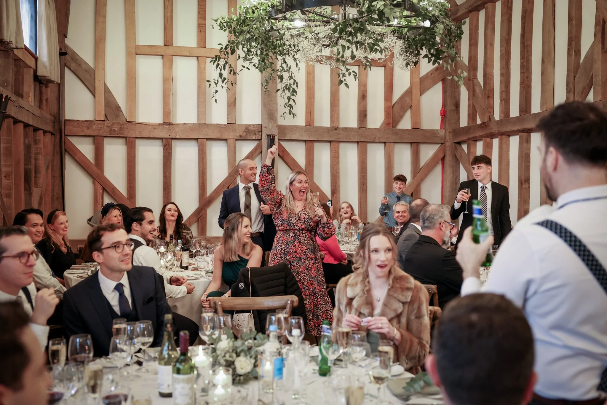 A celebration or wedding reception with guests in formal attire seated at tables in a rustic venue with wooden walls and ceiling beams, some guests are raising glasses and smiling, while a woman in a floral dress is standing and laughing.