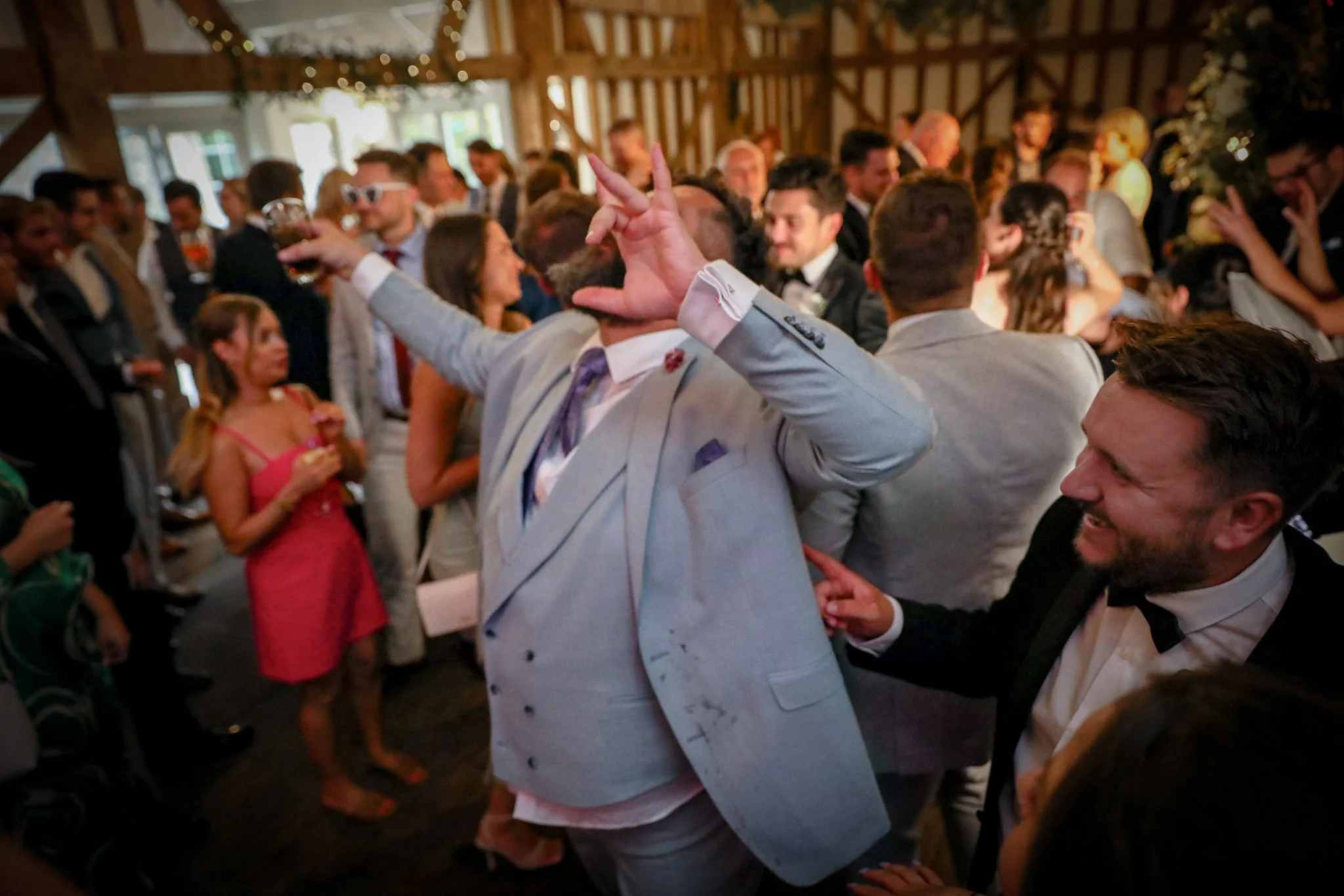 People dancing and socializing at a lively indoor event or party, with a focus on a person in a light-colored suit making a peace sign.