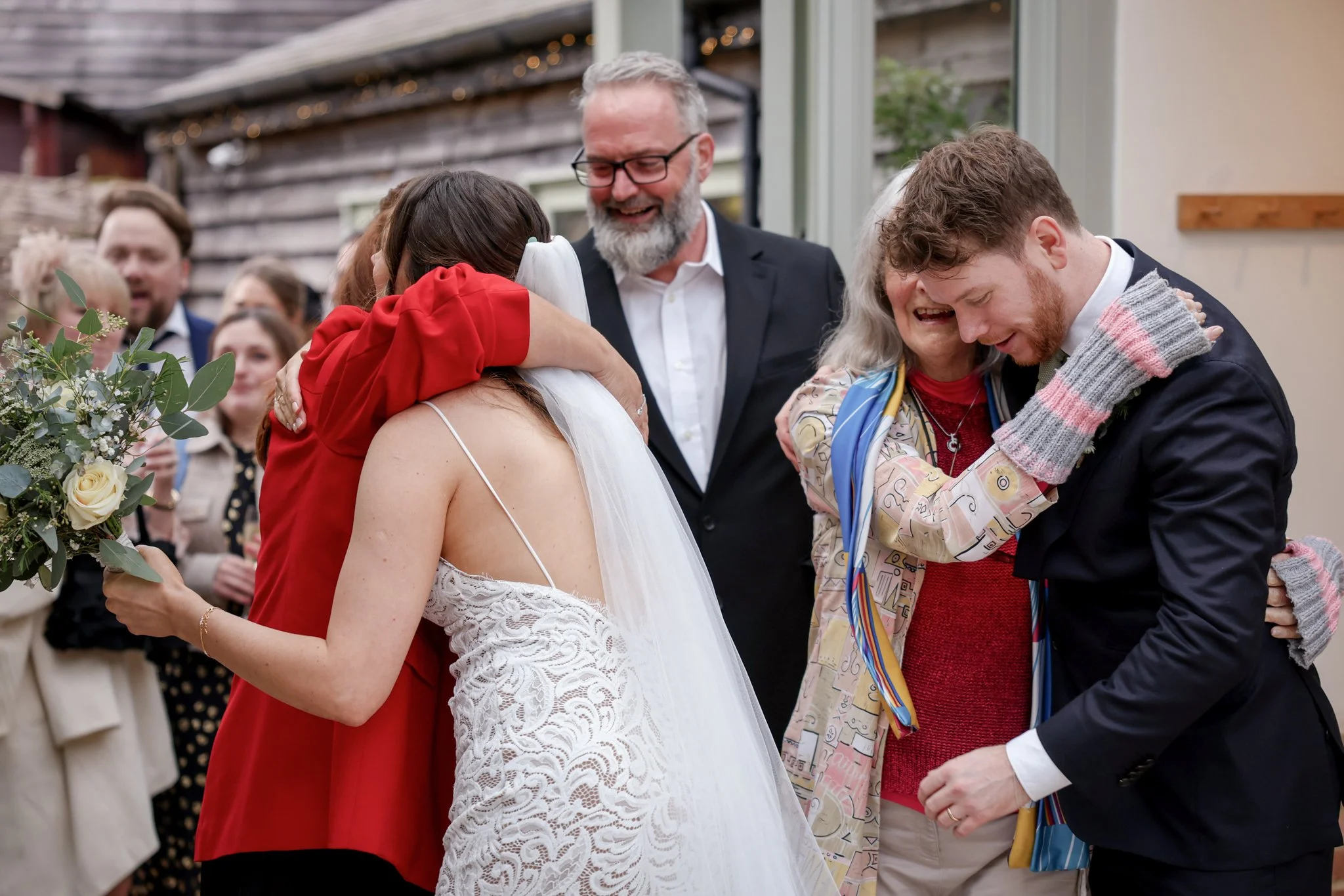 A bride in a white lace dress and veil hugging a woman in a red jacket at a wedding reception, with multiple guests in the background, some embracing and smiling.