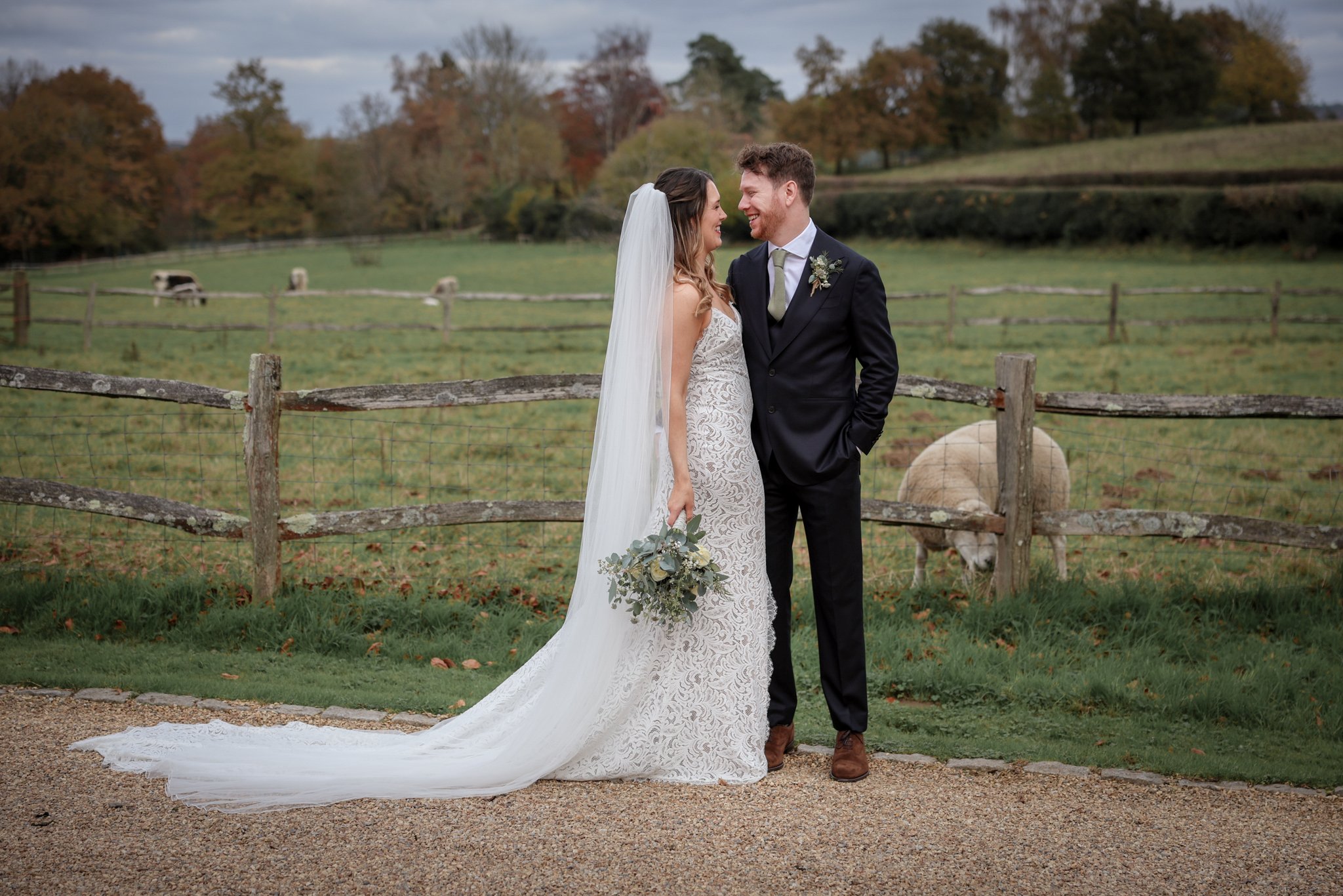 A bride and groom stand close together outdoors in a grassy area with a wooden fence and trees in the background. The bride wears a white lace wedding gown with a long train and veil, holding a bouquet of flowers. The groom wears a black suit with a 