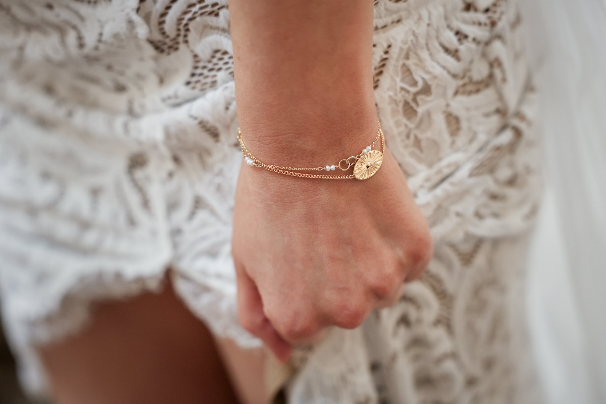 A person's wrist adorned with layered gold bracelets, including one with a circular charm and small pearls, against a textured beige fabric background.
