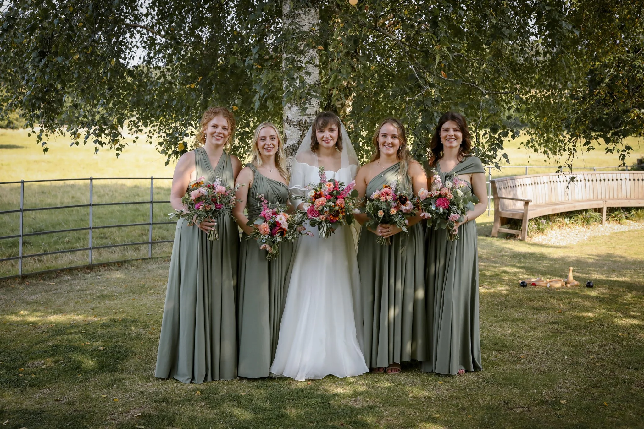 Group of five women in bridesmaid dresses and one bride in a white wedding dress standing outdoors under a tree, holding bouquets of flowers, with a wooden bench and outdoor game equipment on the grass behind them.