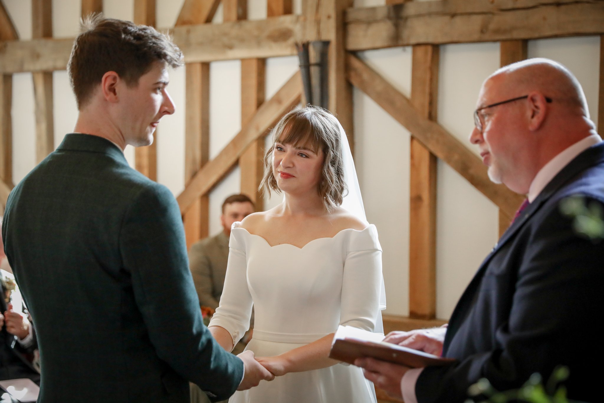 A bride and groom exchanging wedding vows during a ceremony in a rustic barn, with a woman in a white dress overseeing the event and a man in a suit holding a book.