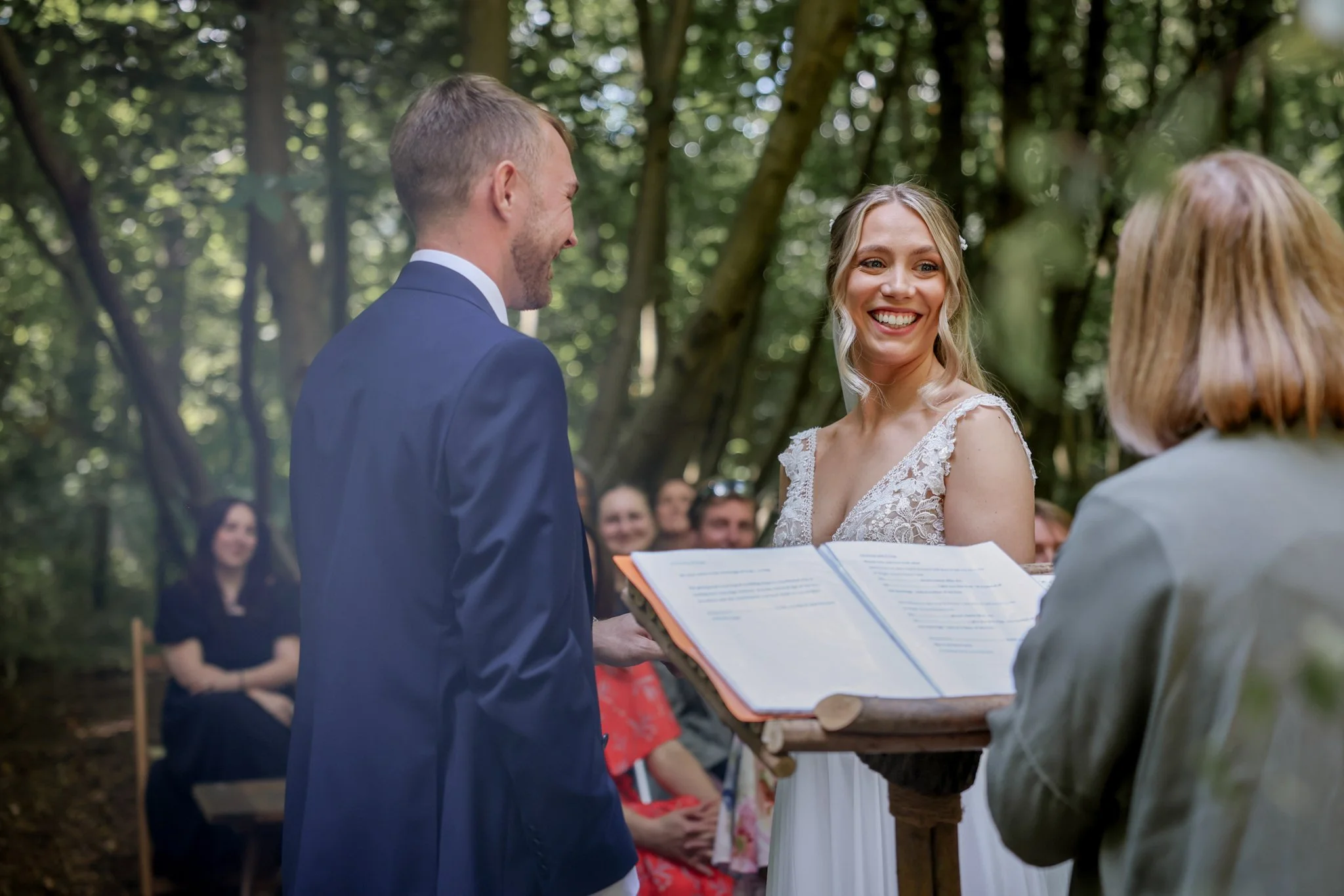 A bride and groom exchanging vows at an outdoor wedding ceremony in a wooded area, with guests in the background.