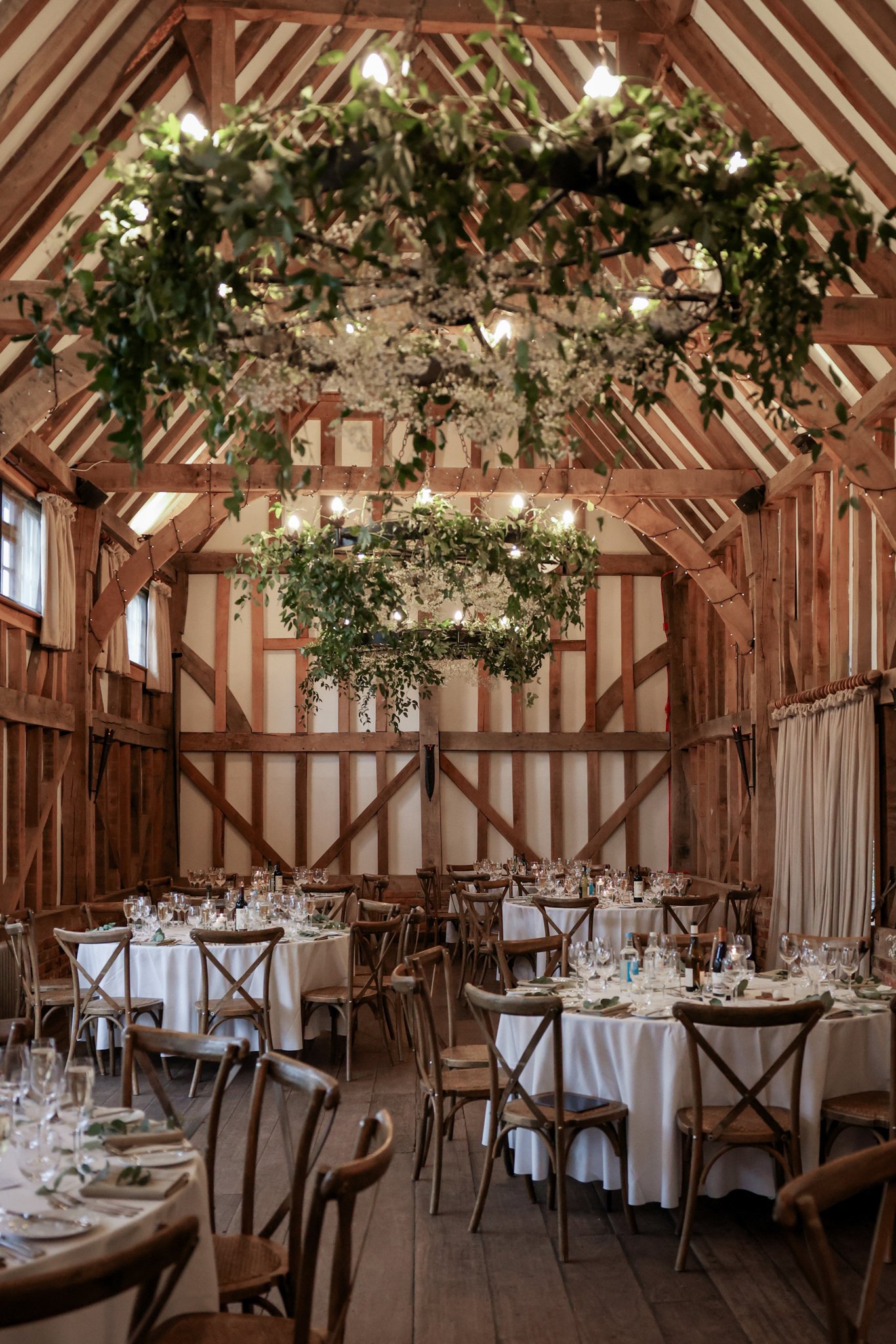 Elegant rustic wedding reception with wooden beams, round tables with white tablecloths, surrounded by wooden chairs, and decorated with greenery and floral centerpieces, overhead chandeliers and string lights.