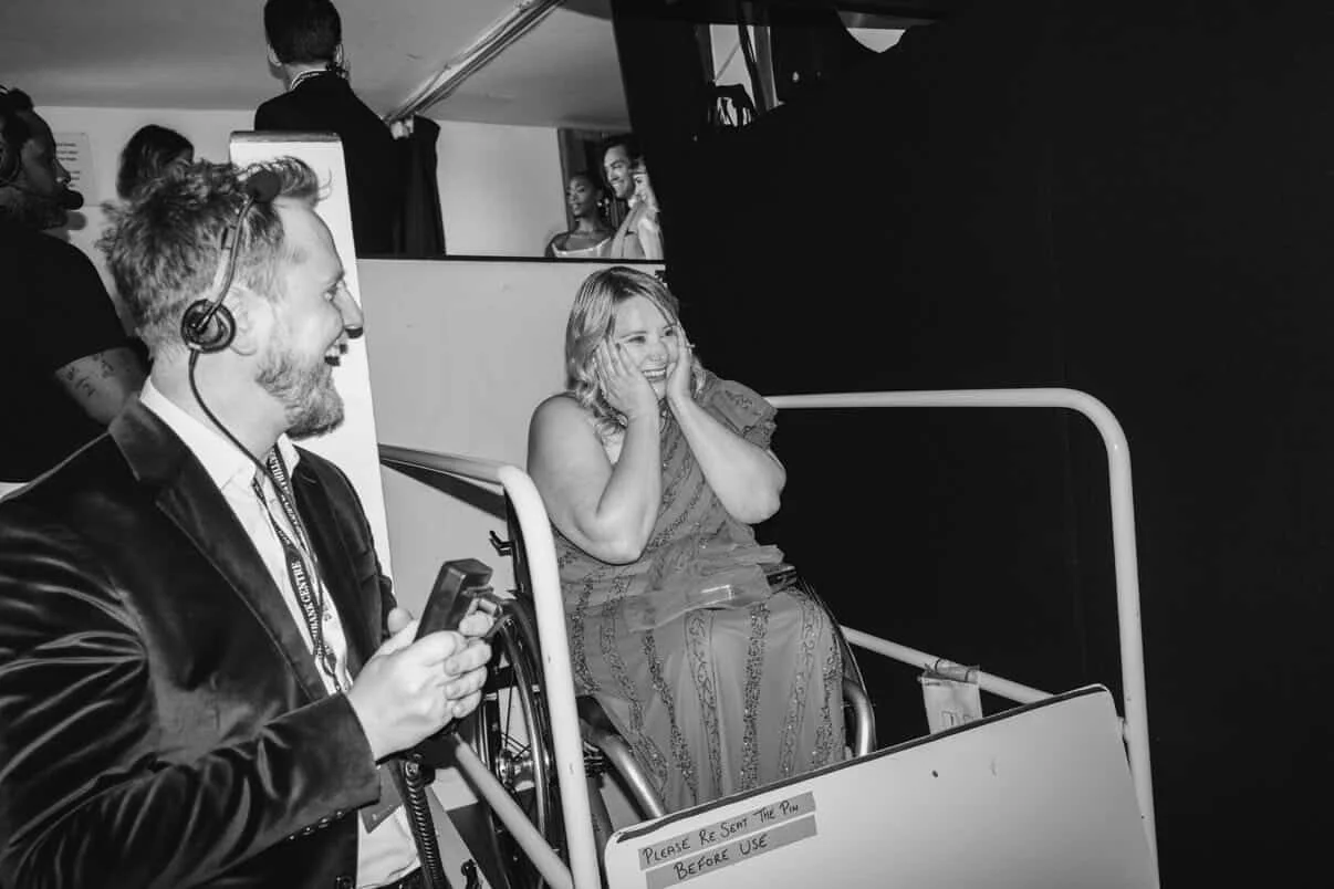 Moritz Valero and Bryony Arnold backstage at the 2026 BAFTA TV Awards. Bryony, a wheelchair user on a stage lift is grabbing her face. Moritz is operating the lift. They both smile face to face.