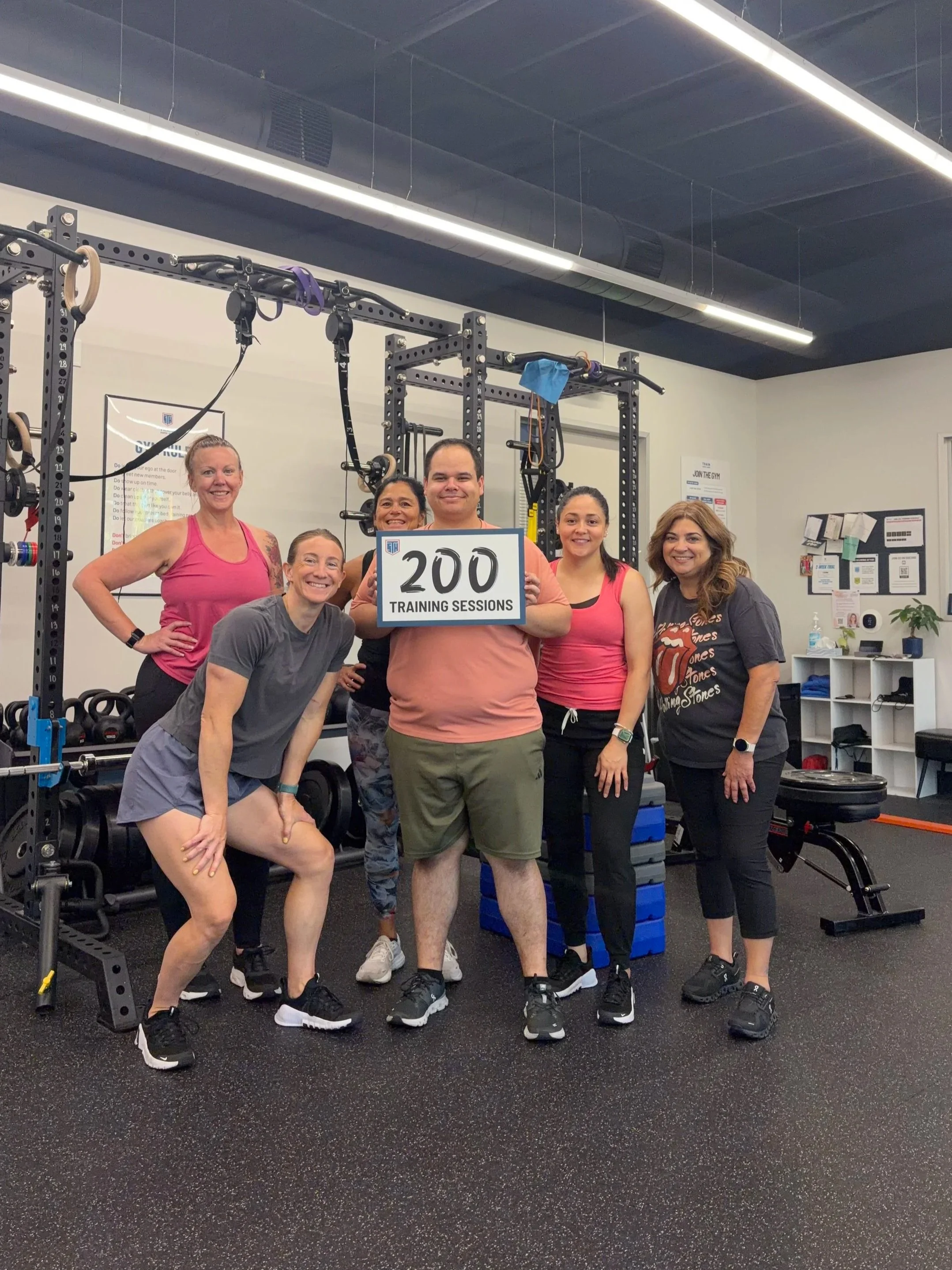 Group of diverse women and men in workout attire in a gym, holding a sign that says "100 Training Sessions" and smiling.