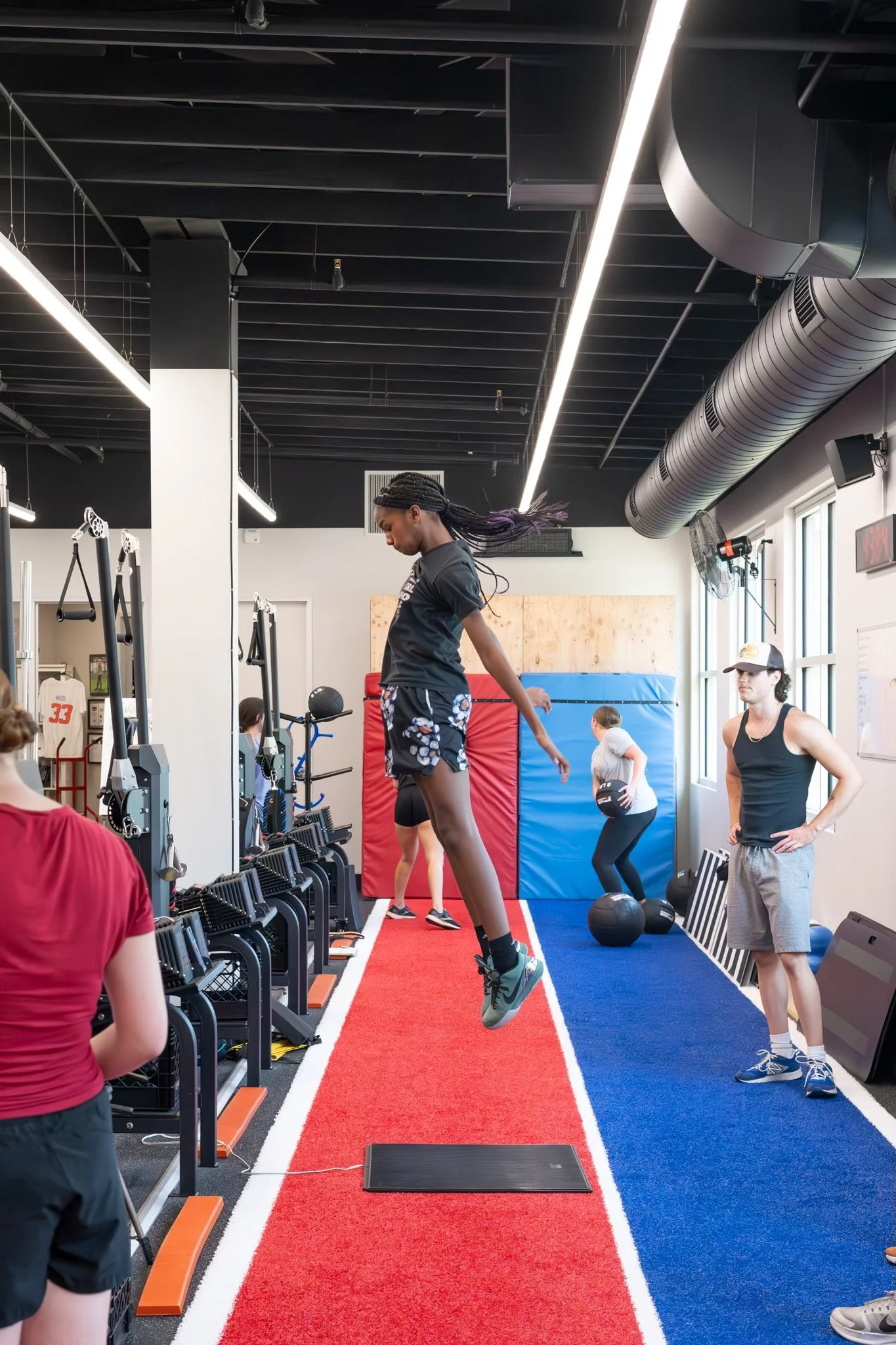 A young man in a white t-shirt and black shorts is lifting weights in a gym. He is standing on a platform in front of a squat rack with black and blue equipment around him.